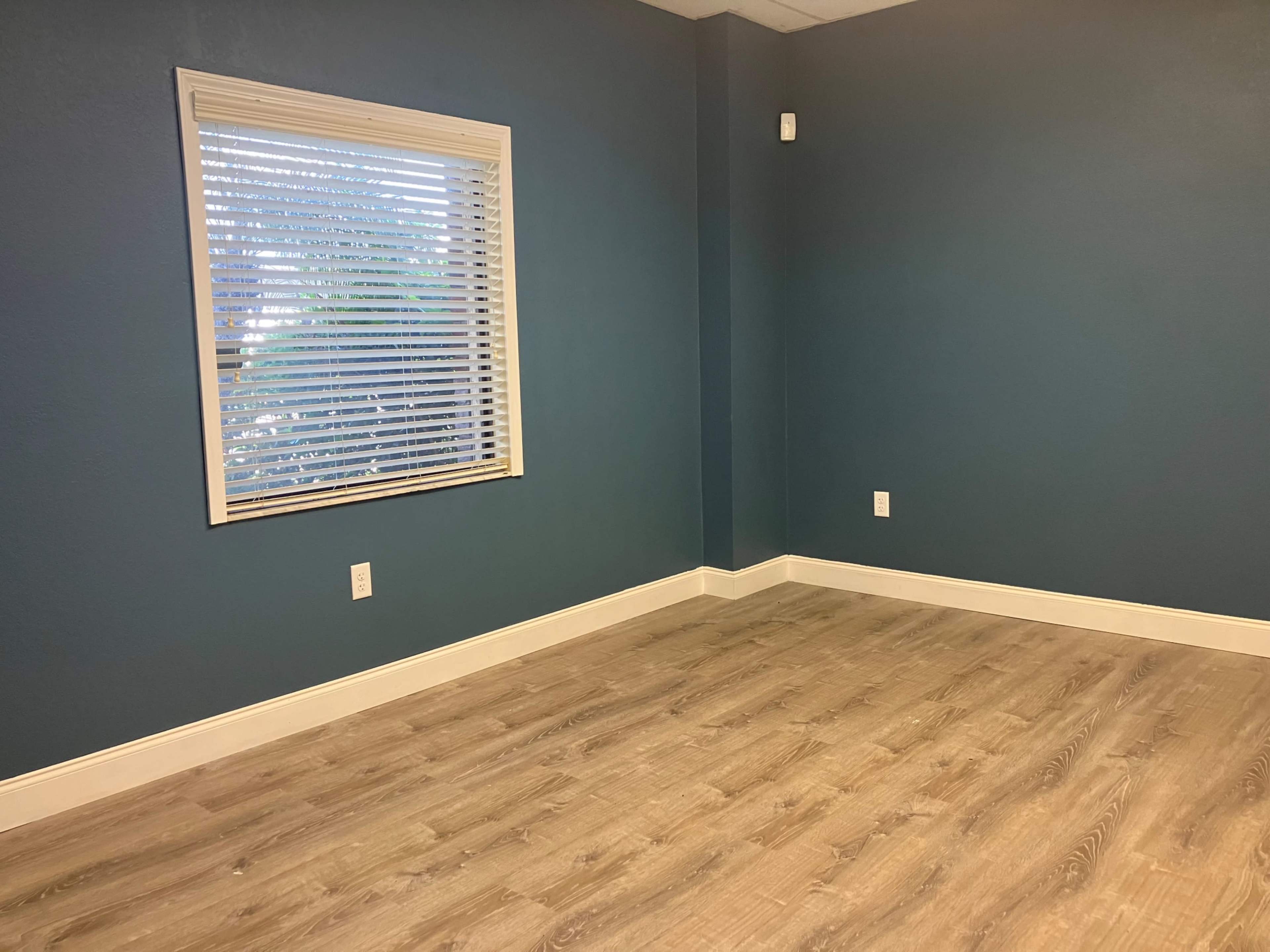A room with blue walls, a window with blinds, and light wooden flooring.