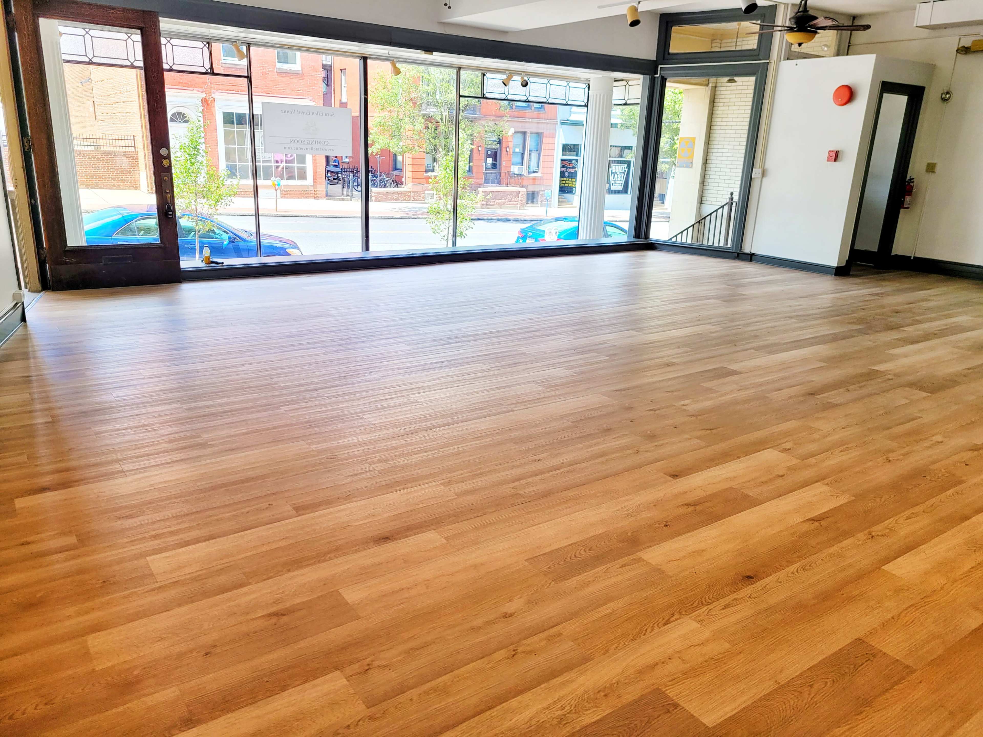 An empty interior space with wooden flooring and large windows facing a street with parked cars and greenery outside.