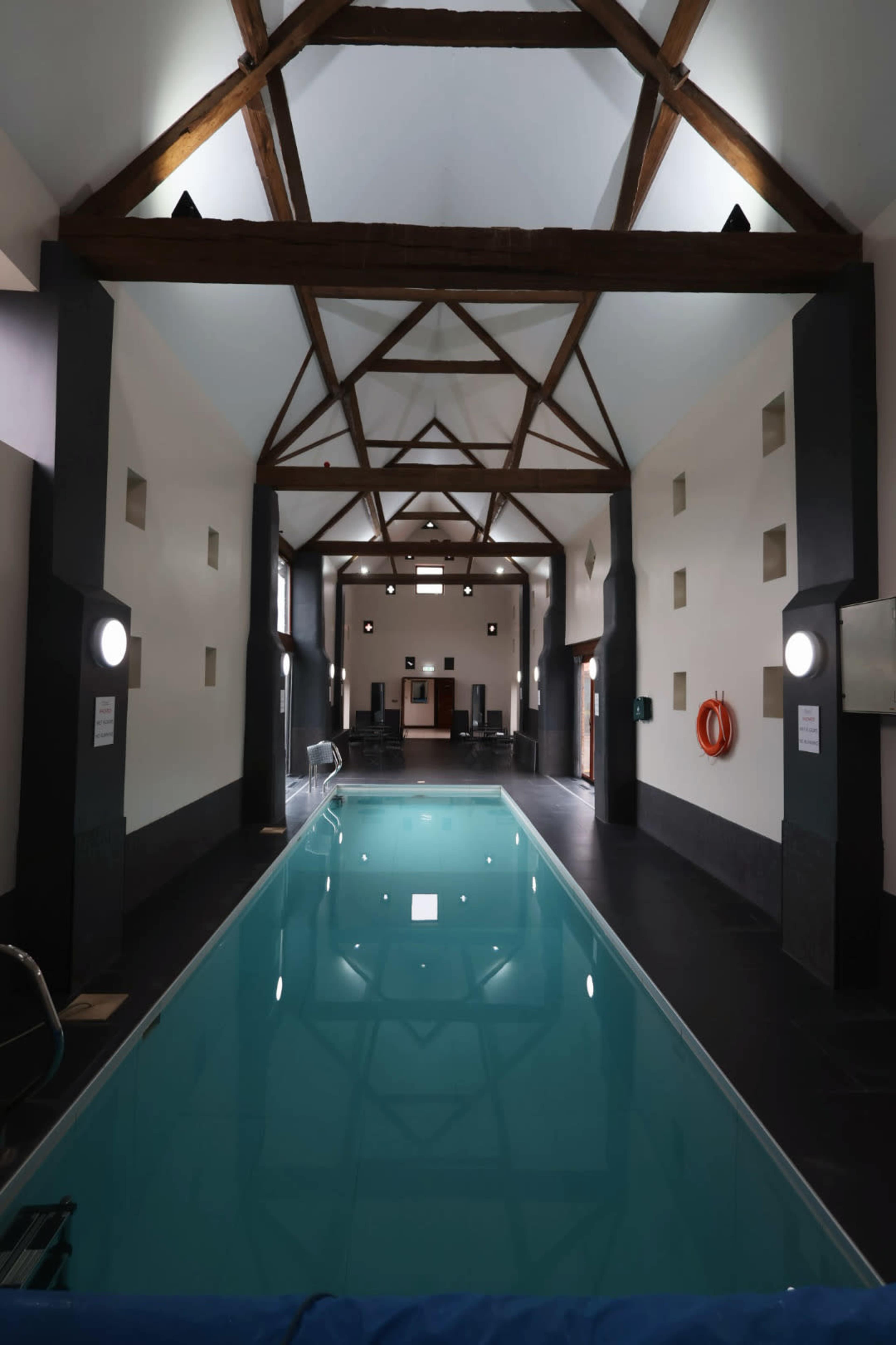 The image shows a spacious indoor swimming pool area with a wooden ceiling, flanked by light-colored walls and a clear blue pool.