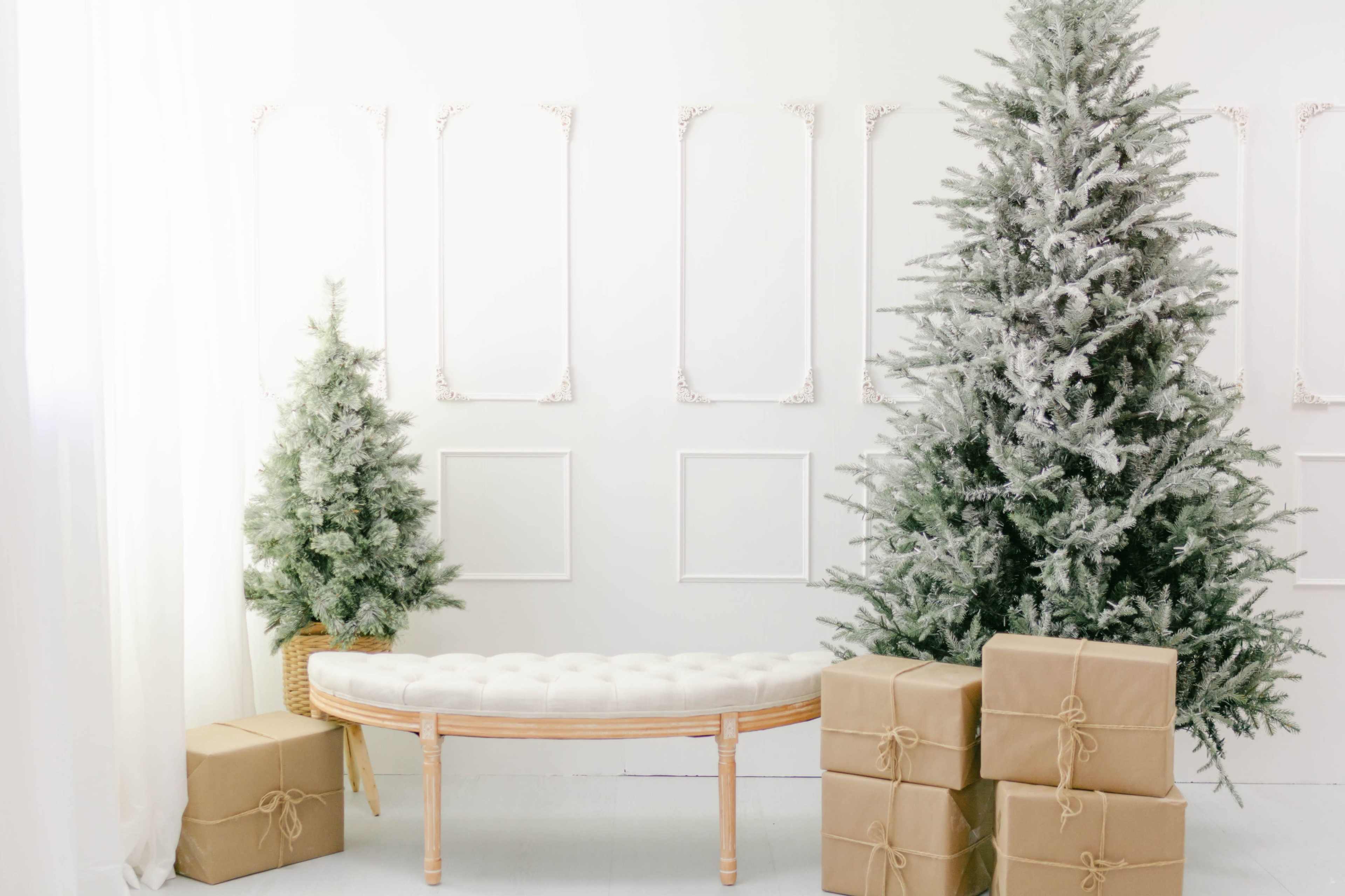 The scene features a decorated room with two Christmas trees, one larger and one smaller, flanking a tufted white bench, accompanied by several wrapped presents in brown paper.