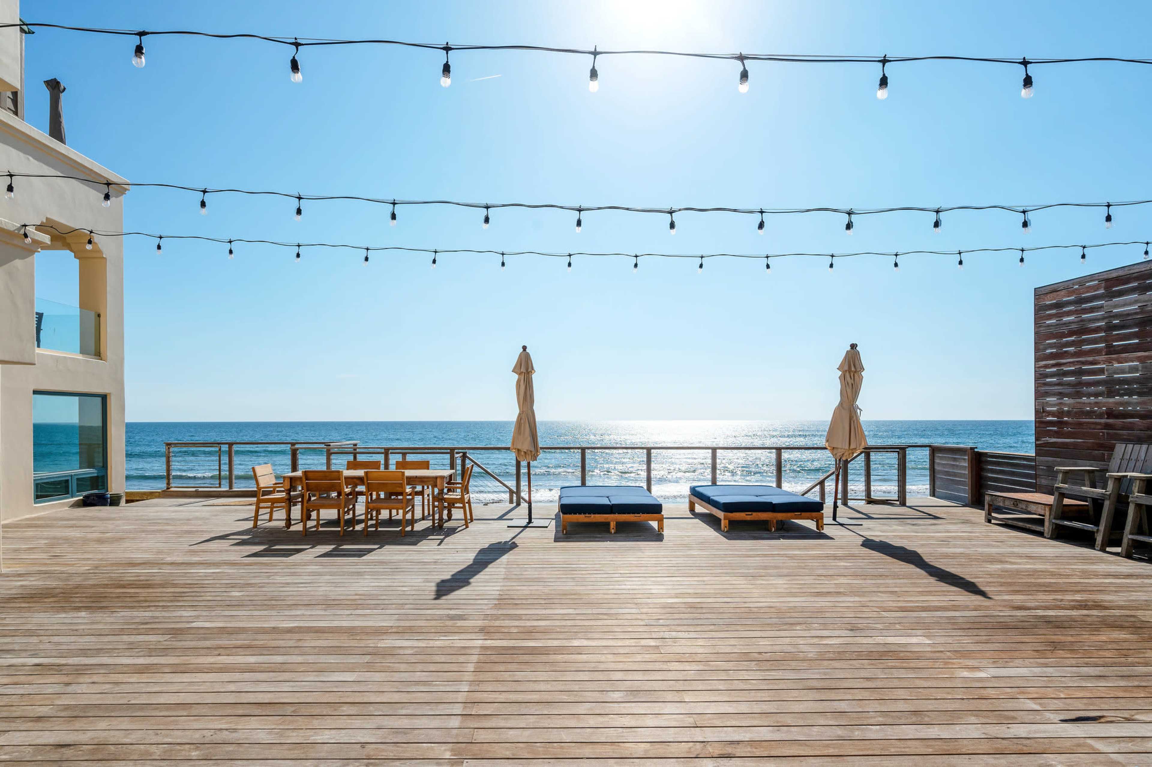 A wooden deck overlooks the ocean, featuring two lounge chairs and a dining area under string lights.
