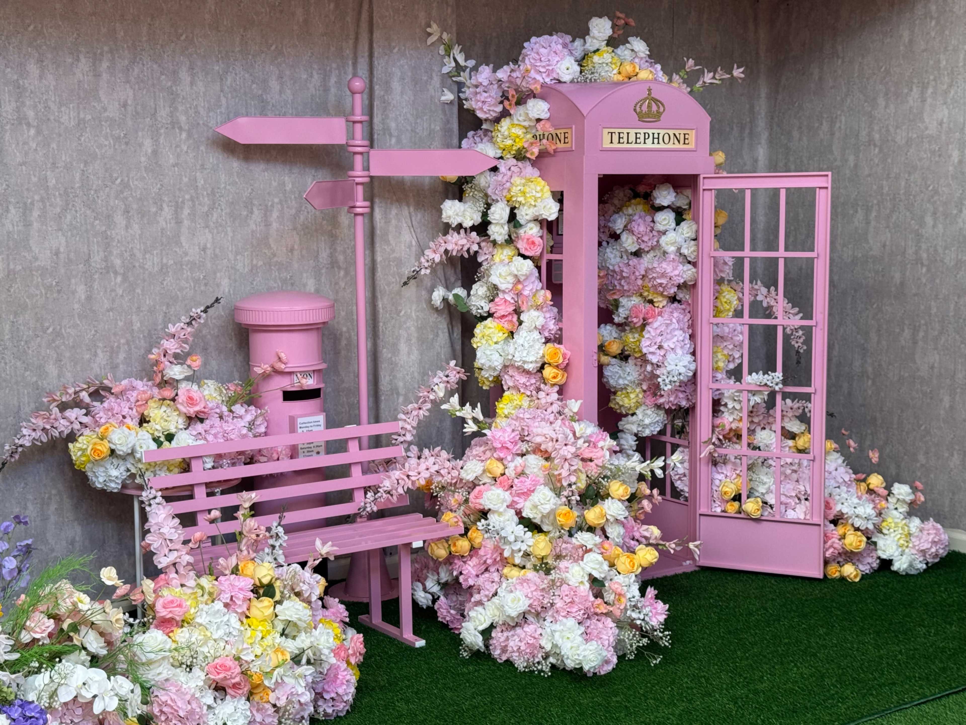 The scene features a pink telephone booth surrounded by colorful artificial flowers, a pink postbox, and a pink bench.
