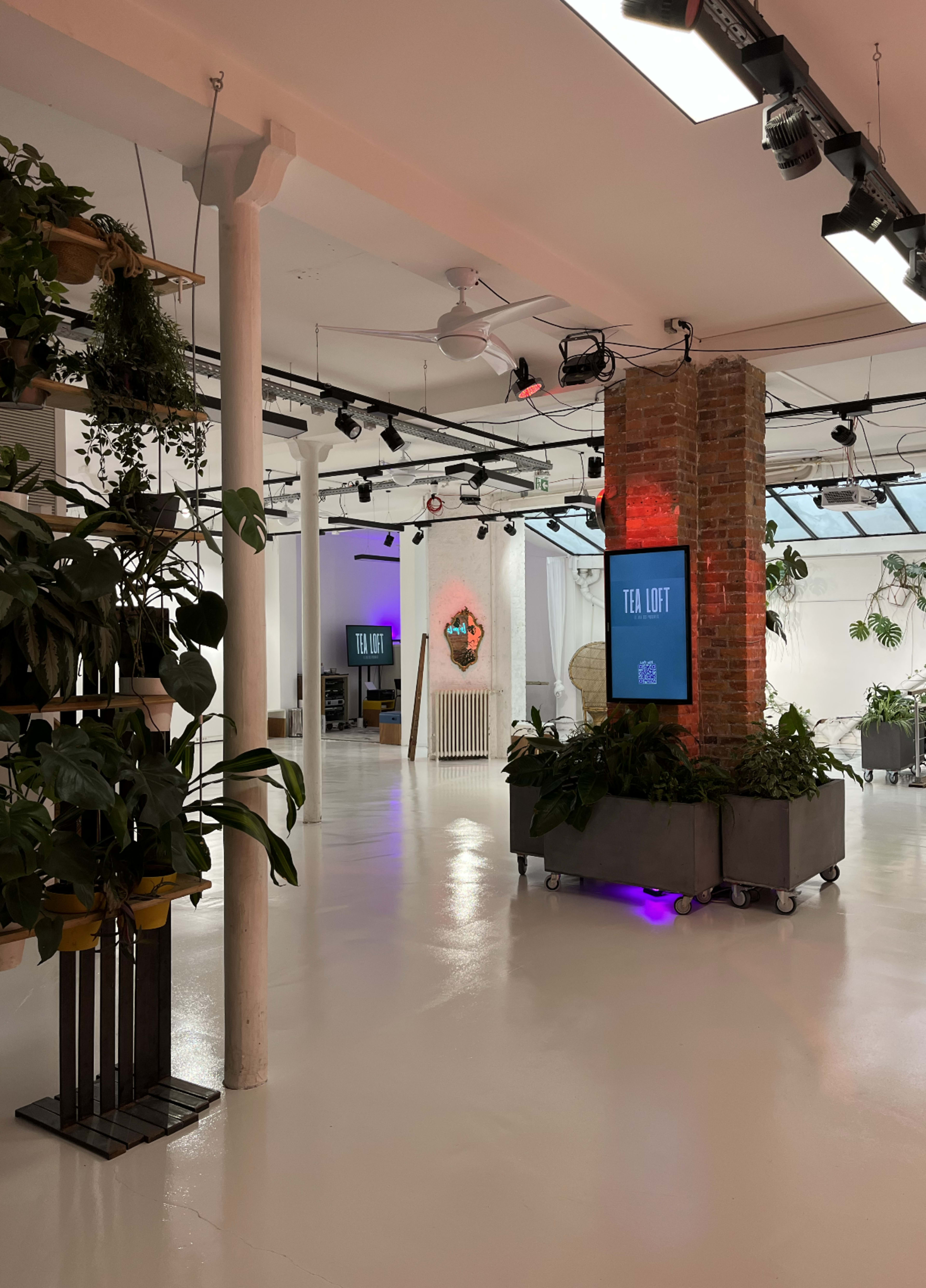 The image shows a modern, well-lit indoor space with large green plants, industrial-style columns, and signage for a "Tea Loft" area.