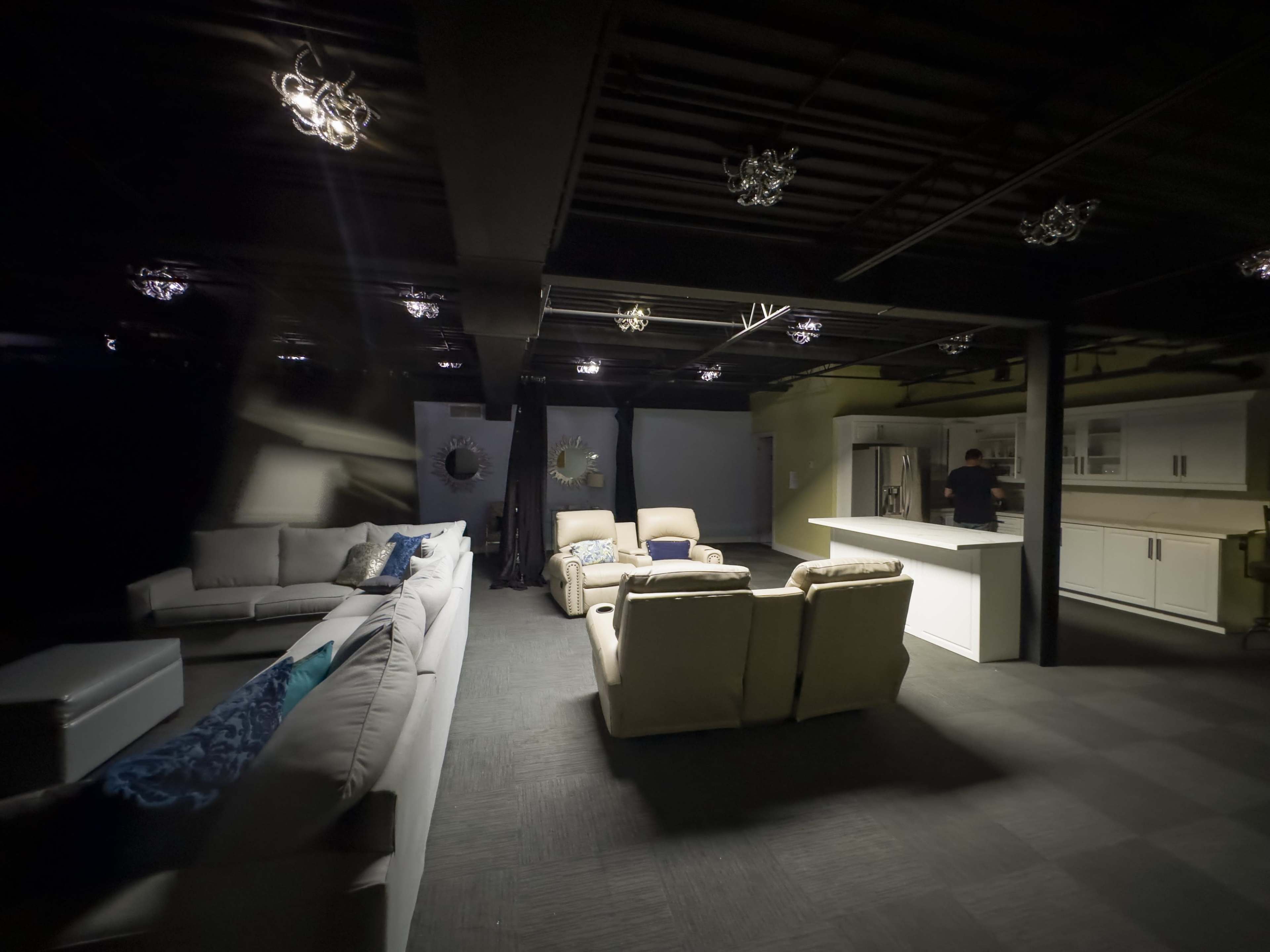 A dimly lit modern living area with two sectional sofas, an island countertop in the background, and a person standing near kitchen appliances.