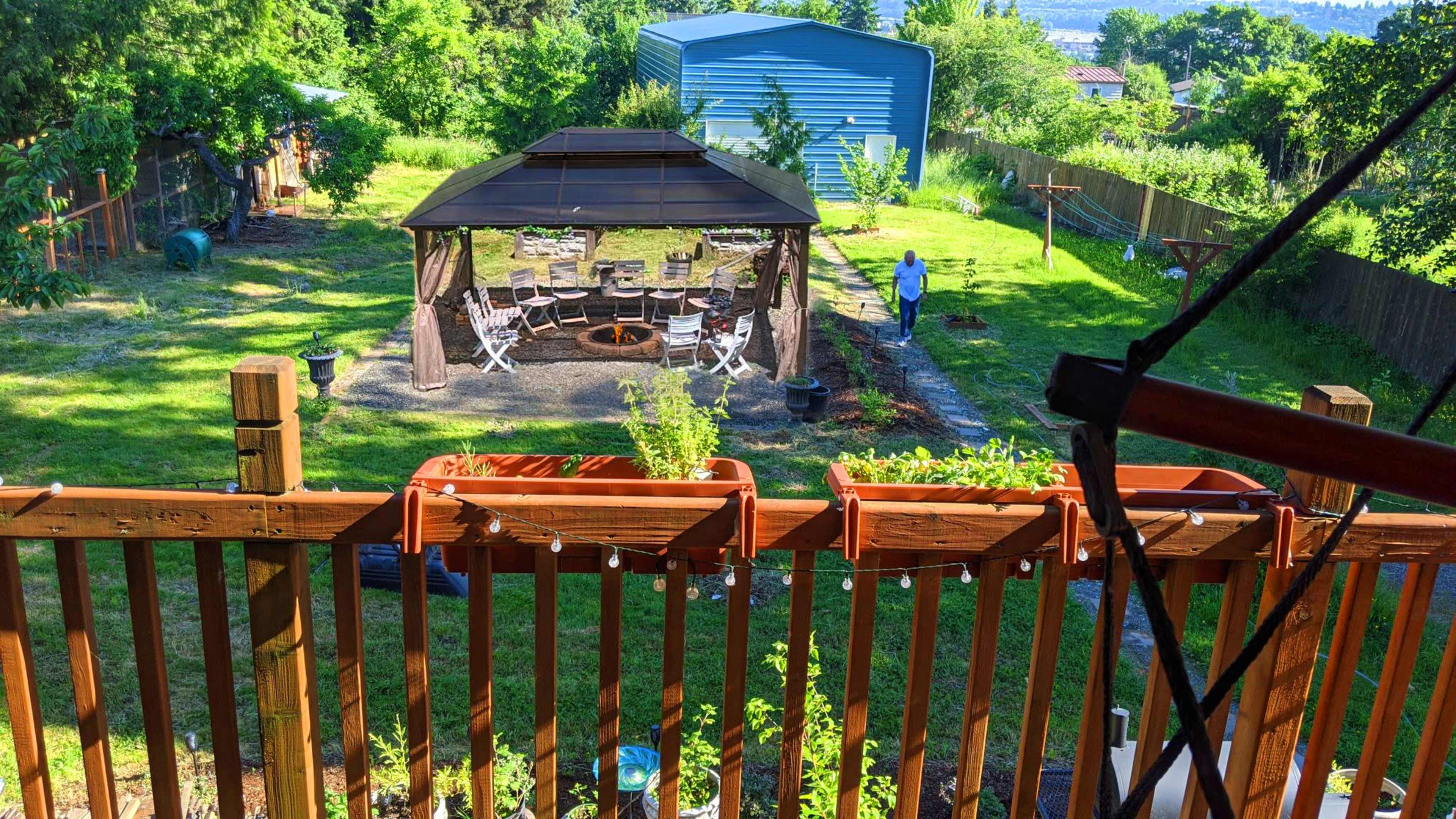 The image shows a backyard with a gazebo, seating area, and a person working in the garden, framed by greenery and a blue shed in the background.