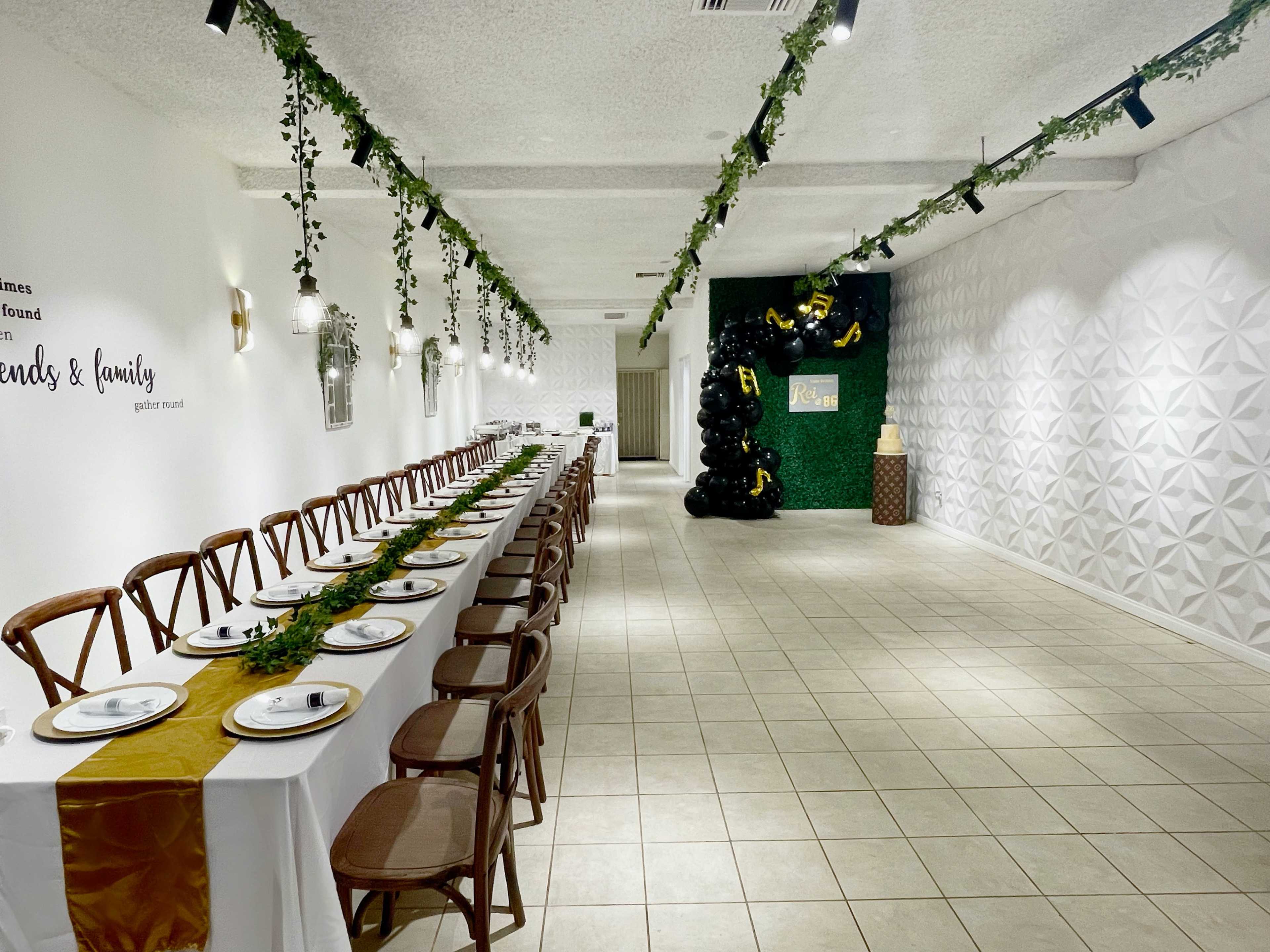 A long, elegantly set dining table runs along the center of a decorated event space with greenery accents and a black balloon display at one end.
