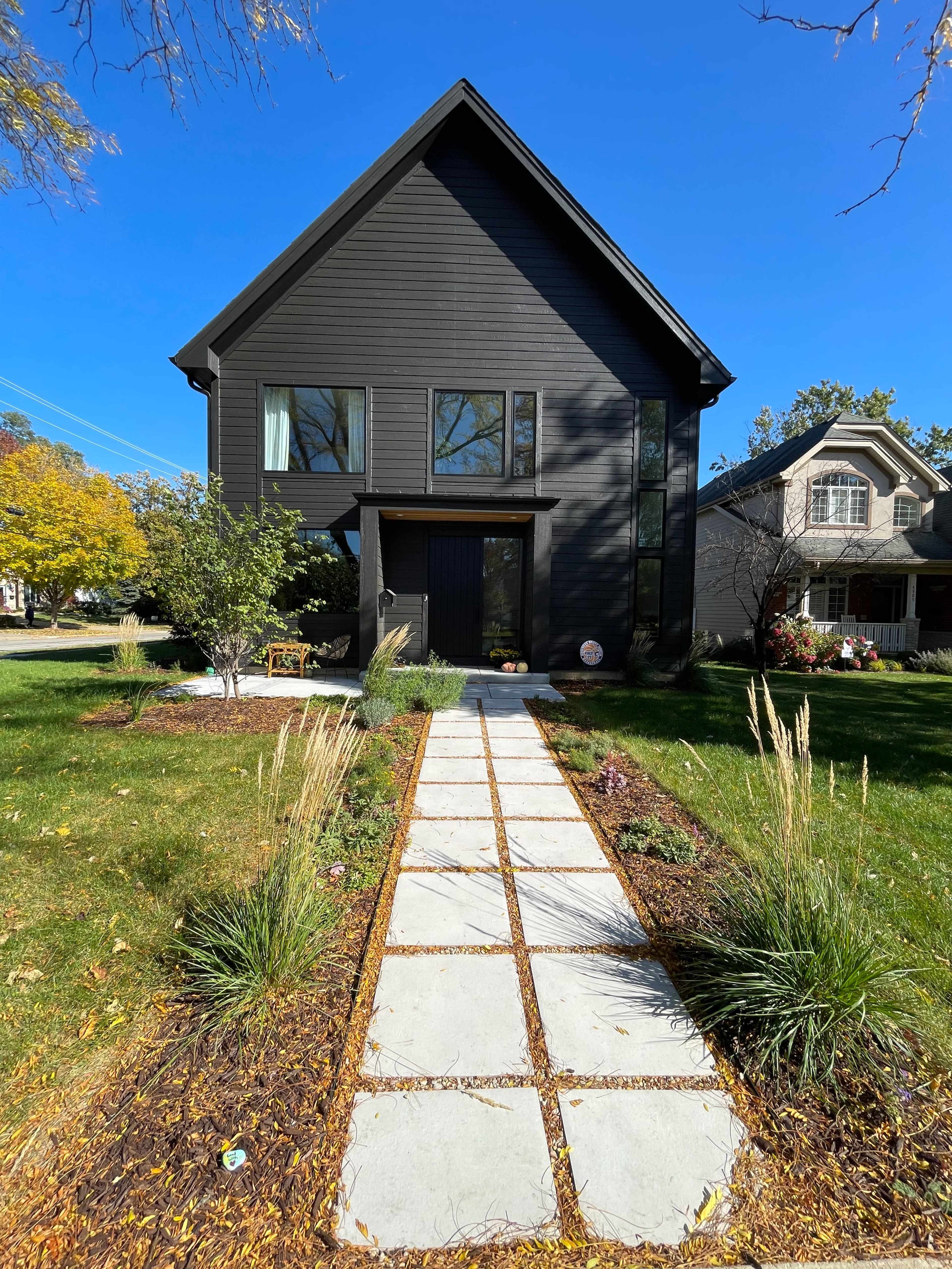 A modern black house stands at the end of a stone pathway lined with grass and shrubs.