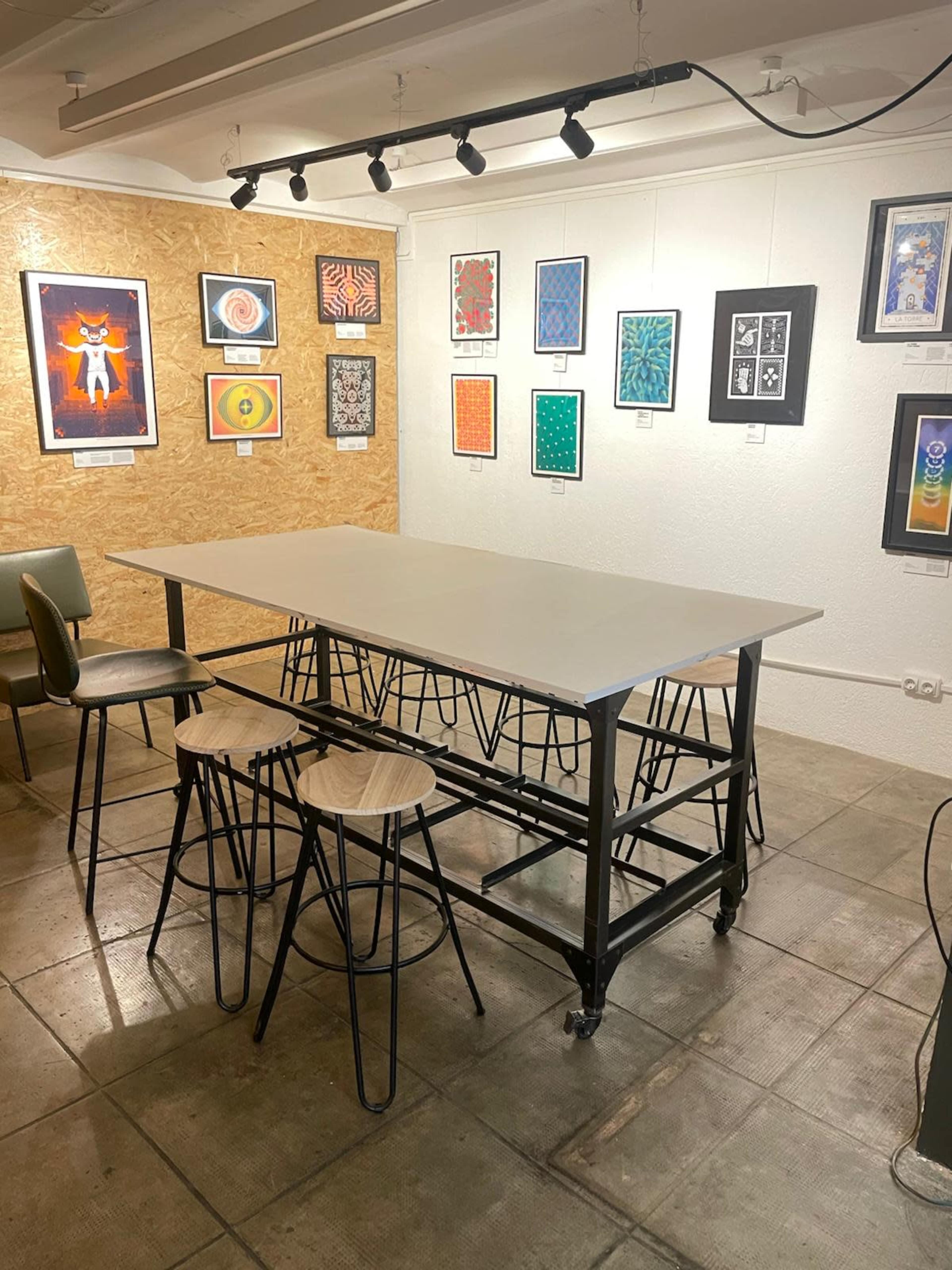 The image shows a modern art gallery featuring a large table with two stools in the foreground and various framed artworks displayed on the walls behind.