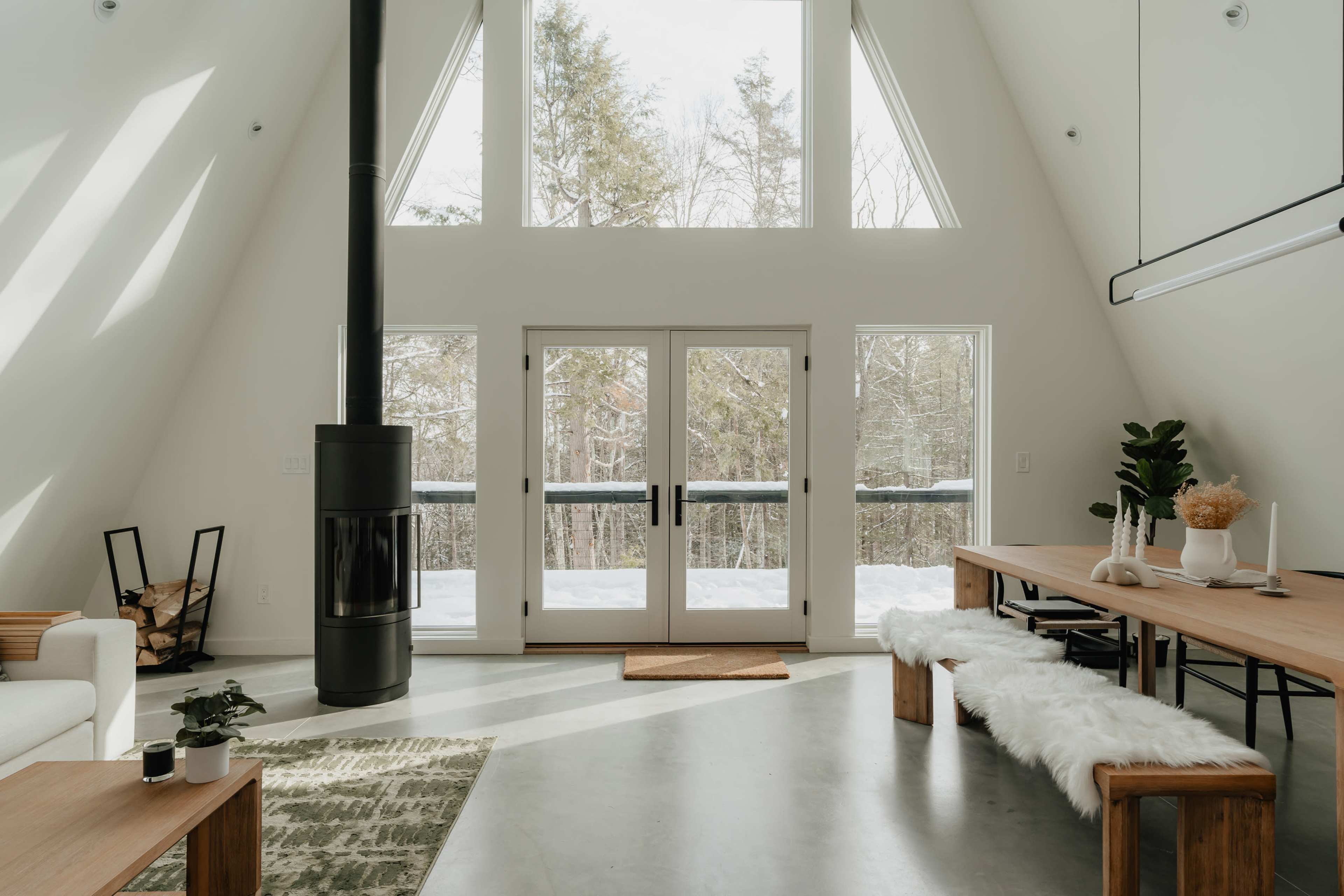 A bright, modern room features large windows, a wooden dining table, and a central wood stove, with snowy trees visible outside.