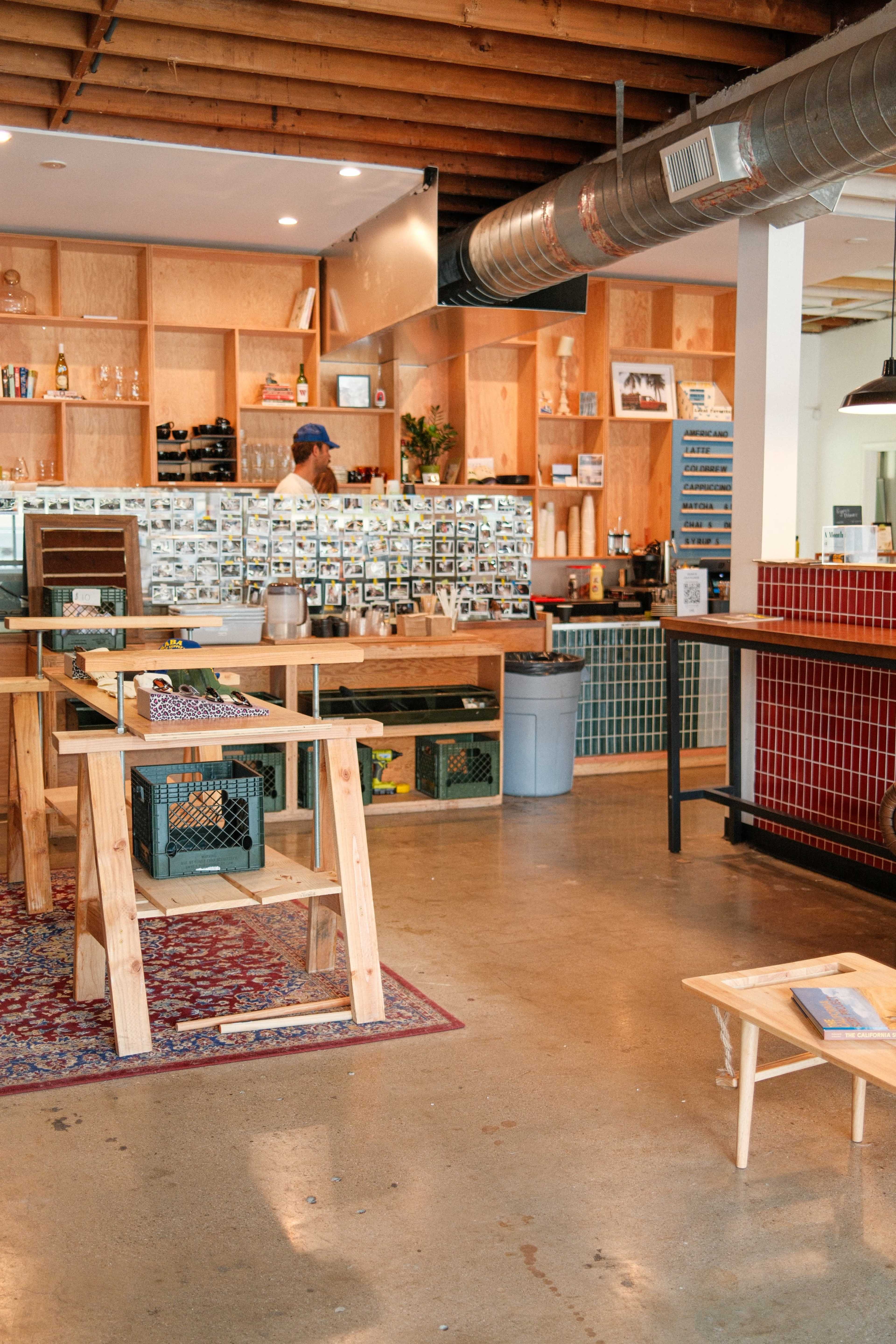 The image shows a modern café interior with wooden furniture, a red tiled counter, and a wall of small photographs behind the barista.
