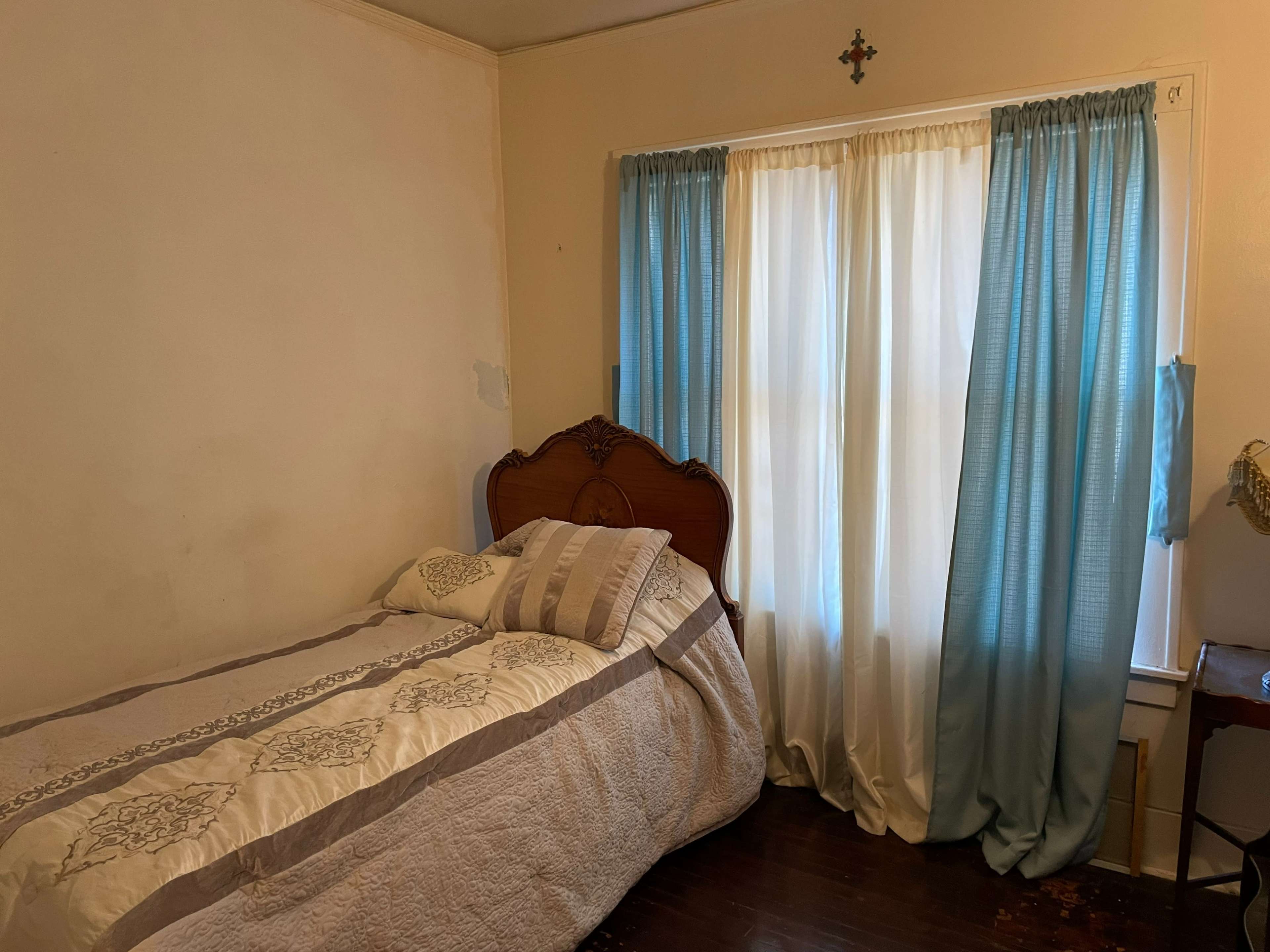 A simple bedroom with a single bed covered in a quilt, a window dressed with light blue curtains, and a small side table.