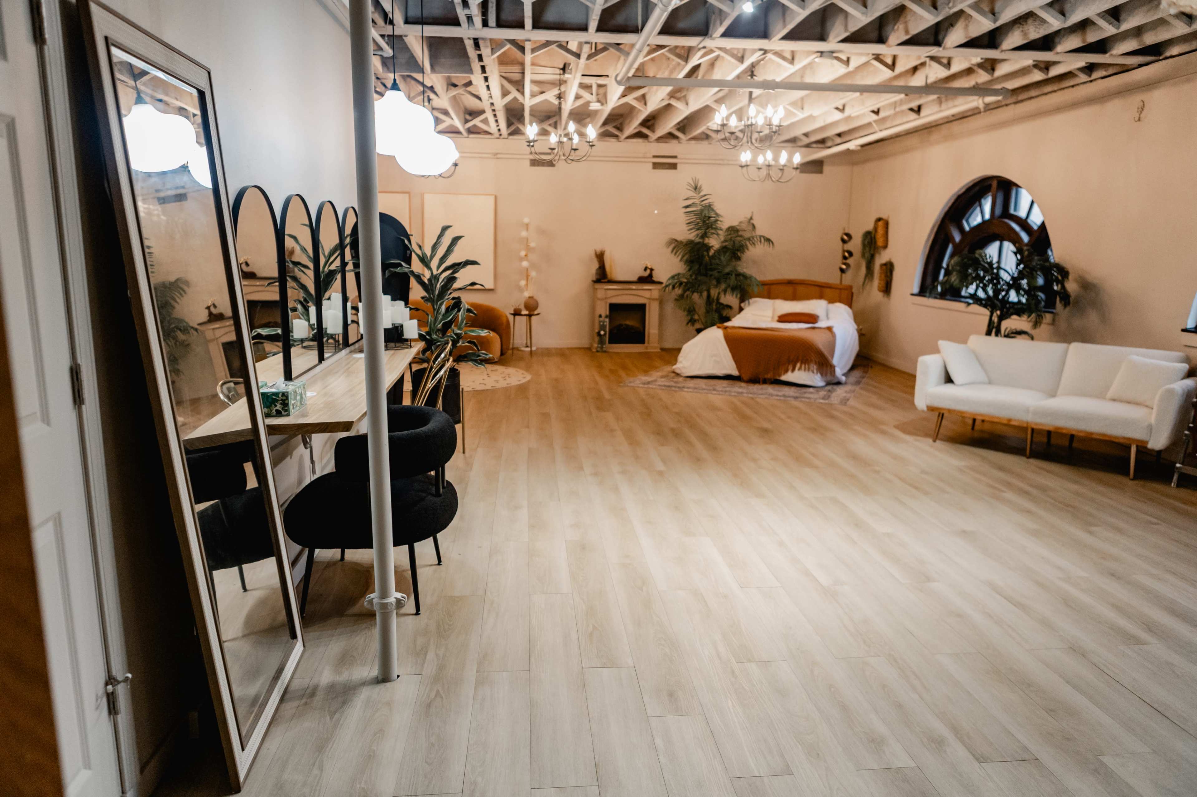 The image shows a spacious room with wooden floors, featuring a bed with brown bedding, a white couch, a large mirror, and various plants.