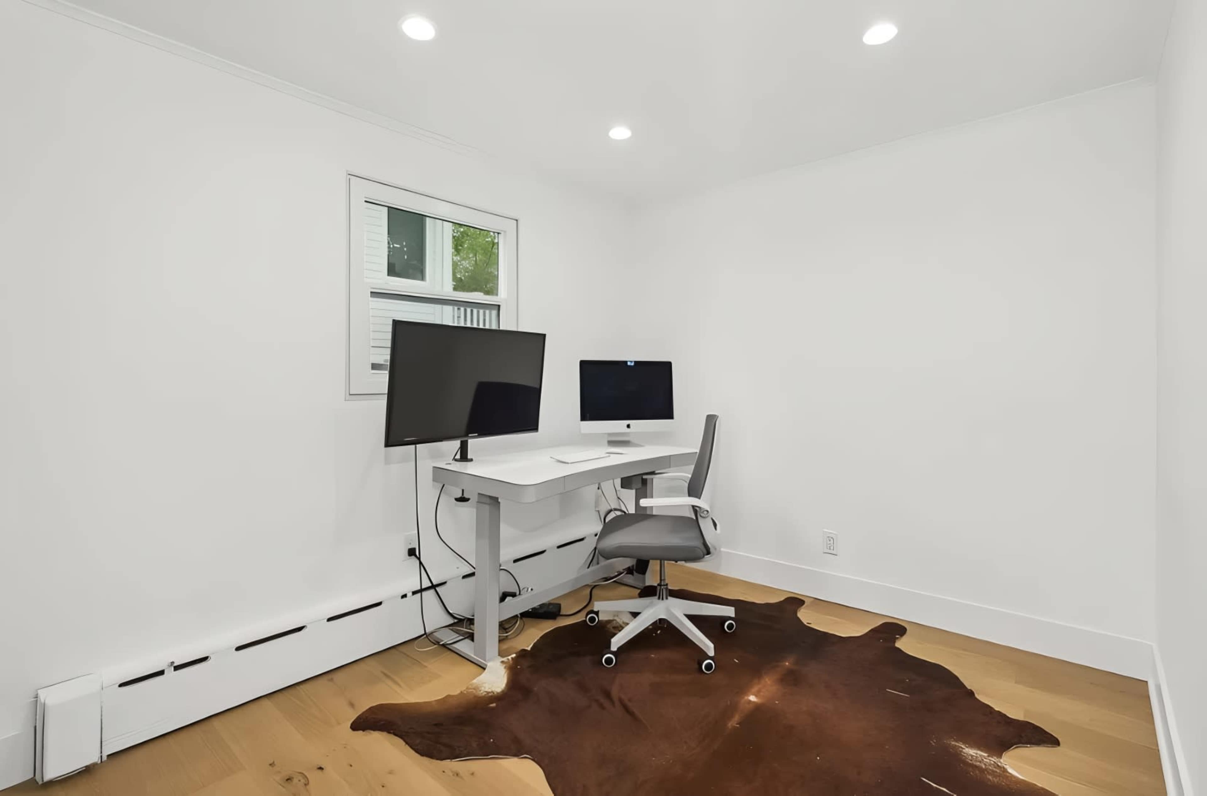 The image shows a minimalist home office with two monitors, a desk, an ergonomic chair, and a cowhide rug on a wooden floor.