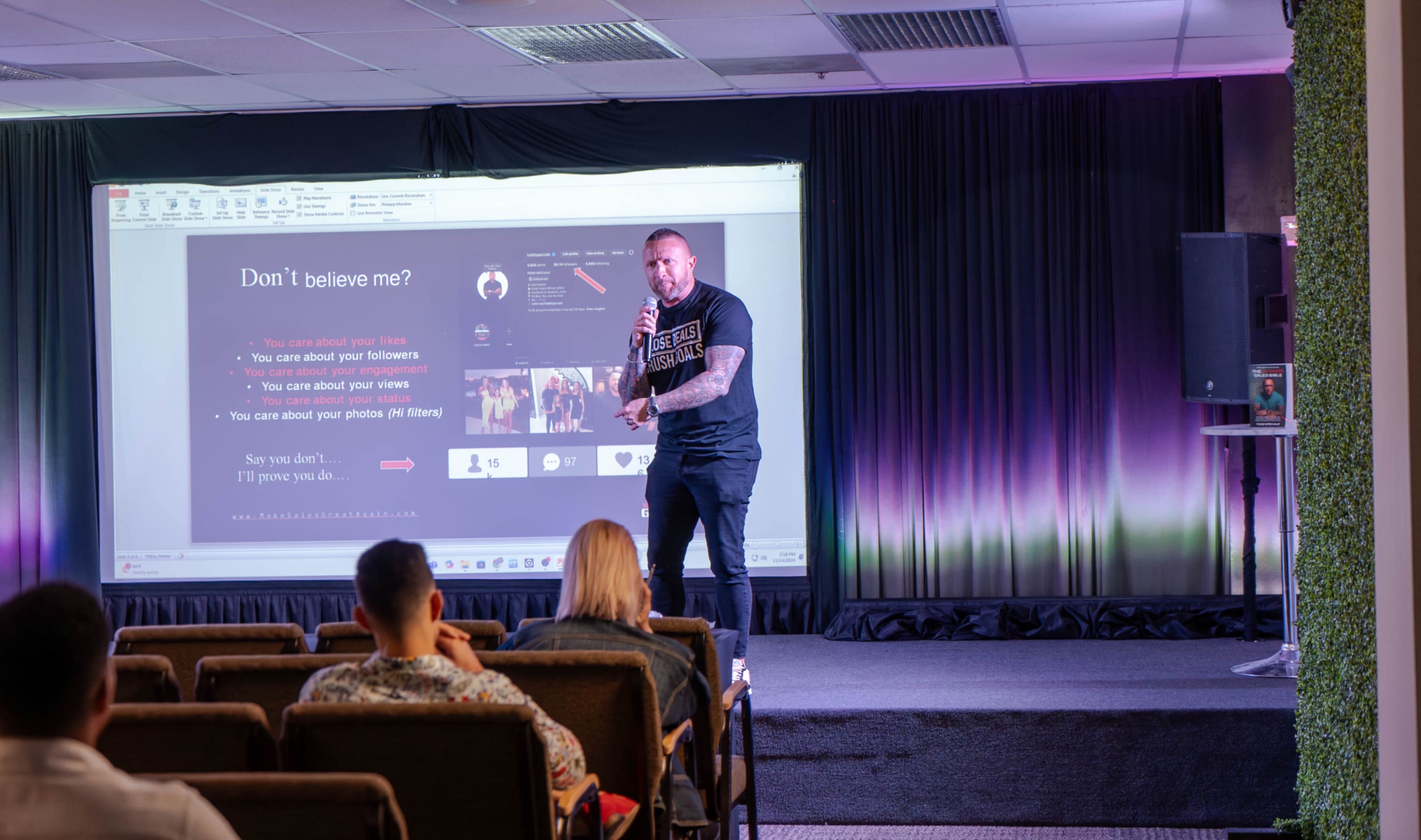 A speaker presents in front of an audience, with a projected slide displaying text and graphics on a large screen.
