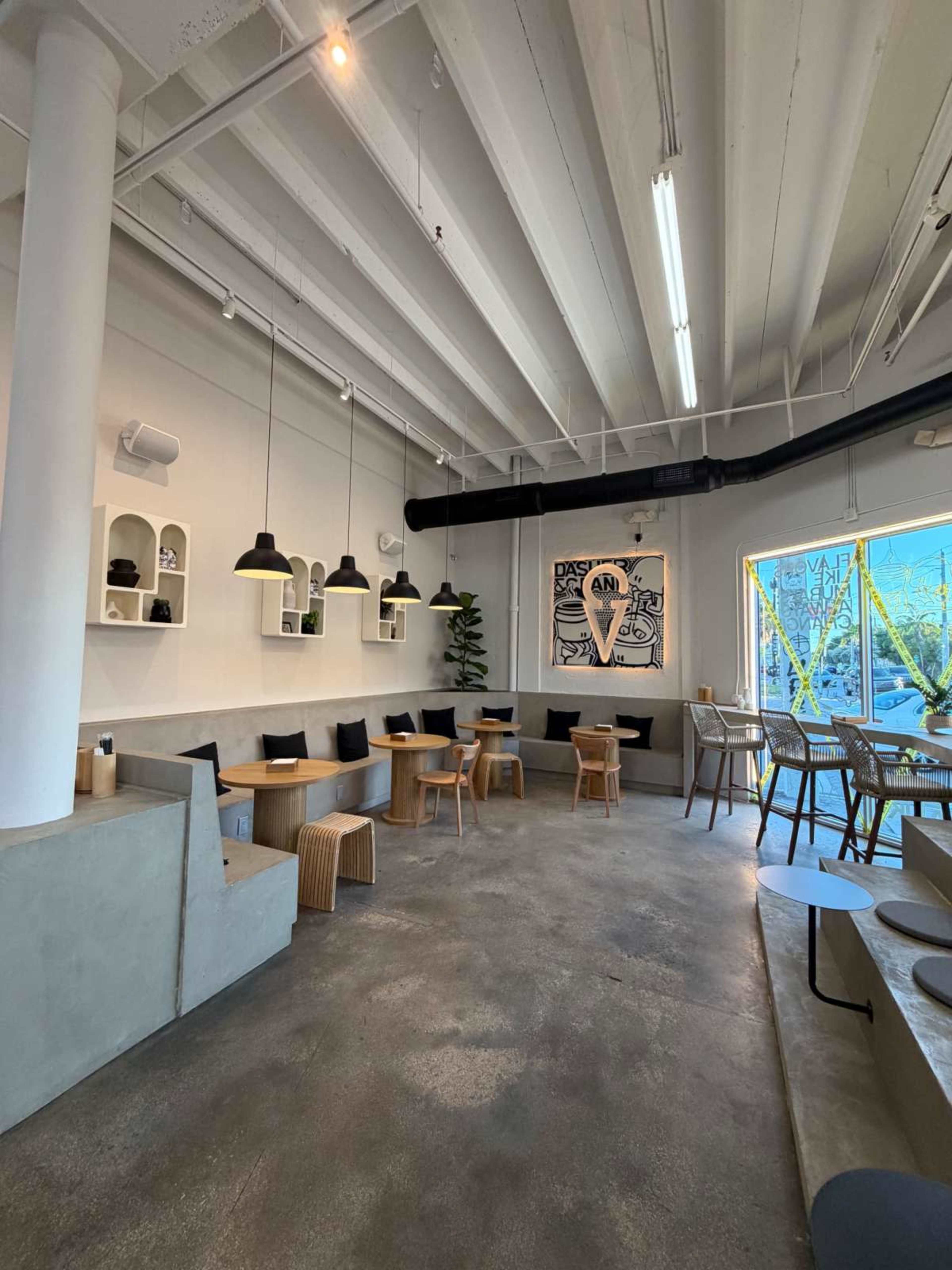The image shows a modern café interior with a mix of seating areas, including wooden tables and chairs, minimalist decor, and large windows letting in natural light.