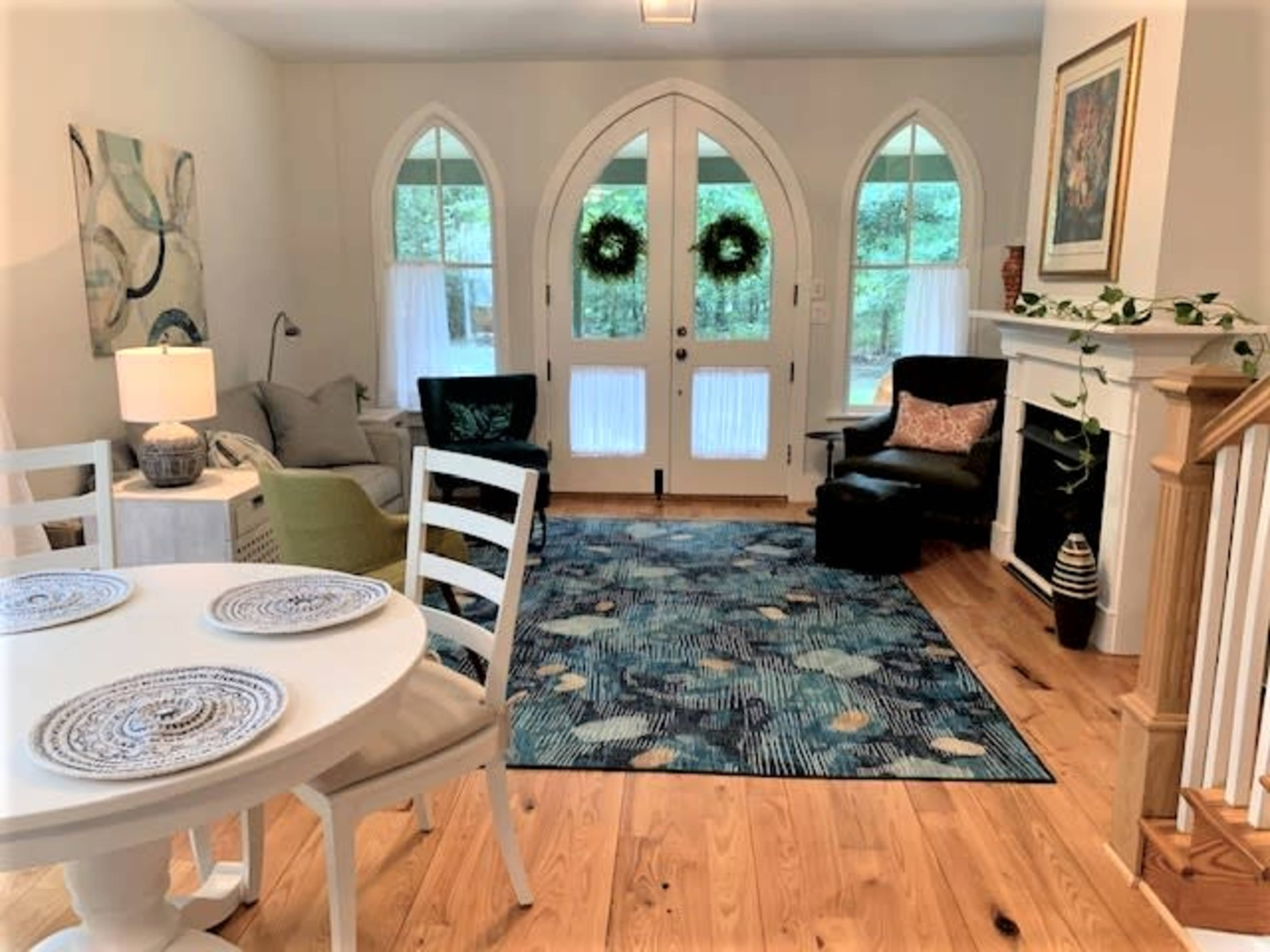A bright living room features a round dining table with white chairs, a green sofa, a black armchair, and large arched windows adorned with decorative wreaths.