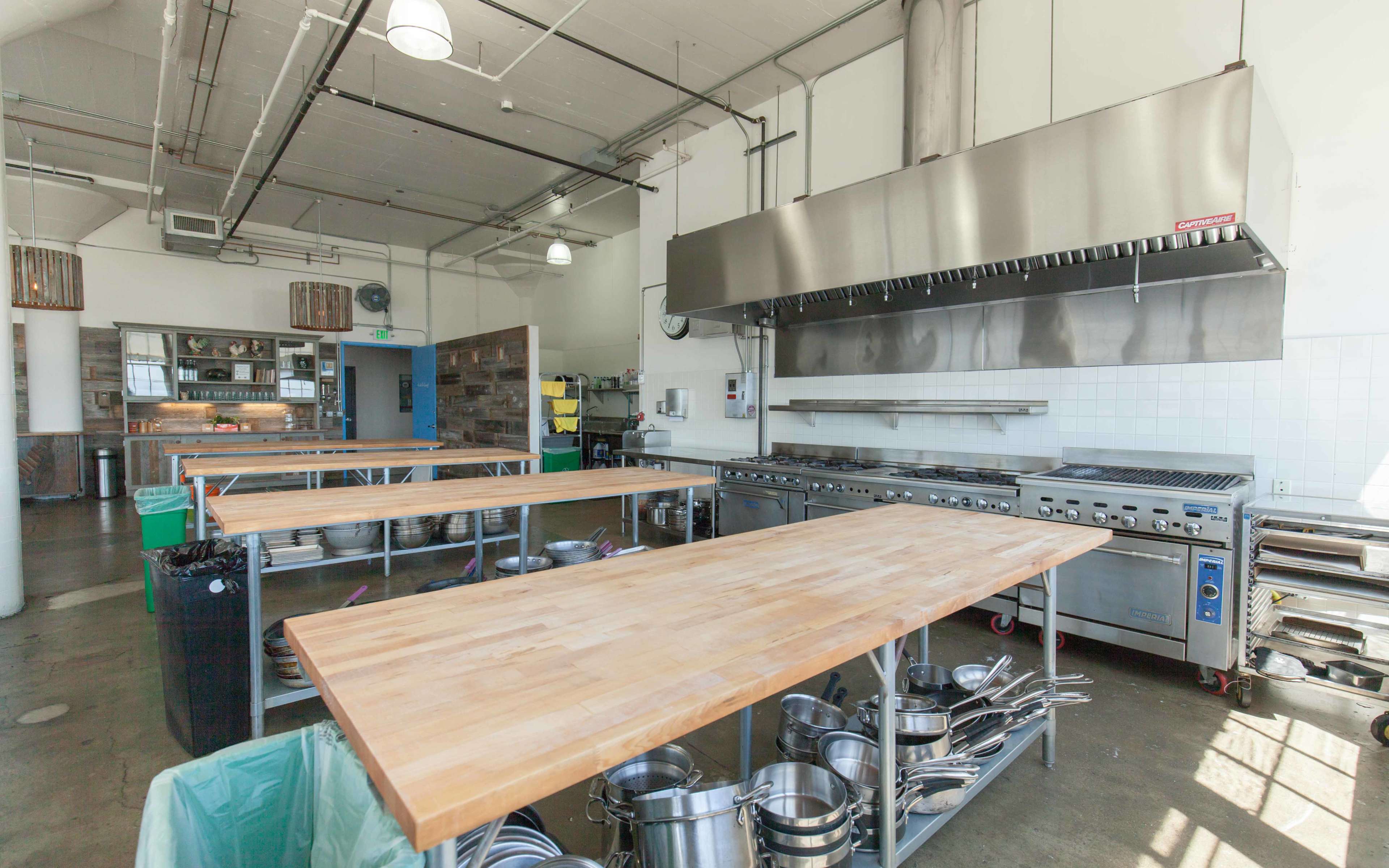 A spacious commercial kitchen features multiple wooden prep tables, a large stainless steel cooking range, and various kitchen utensils organized nearby.