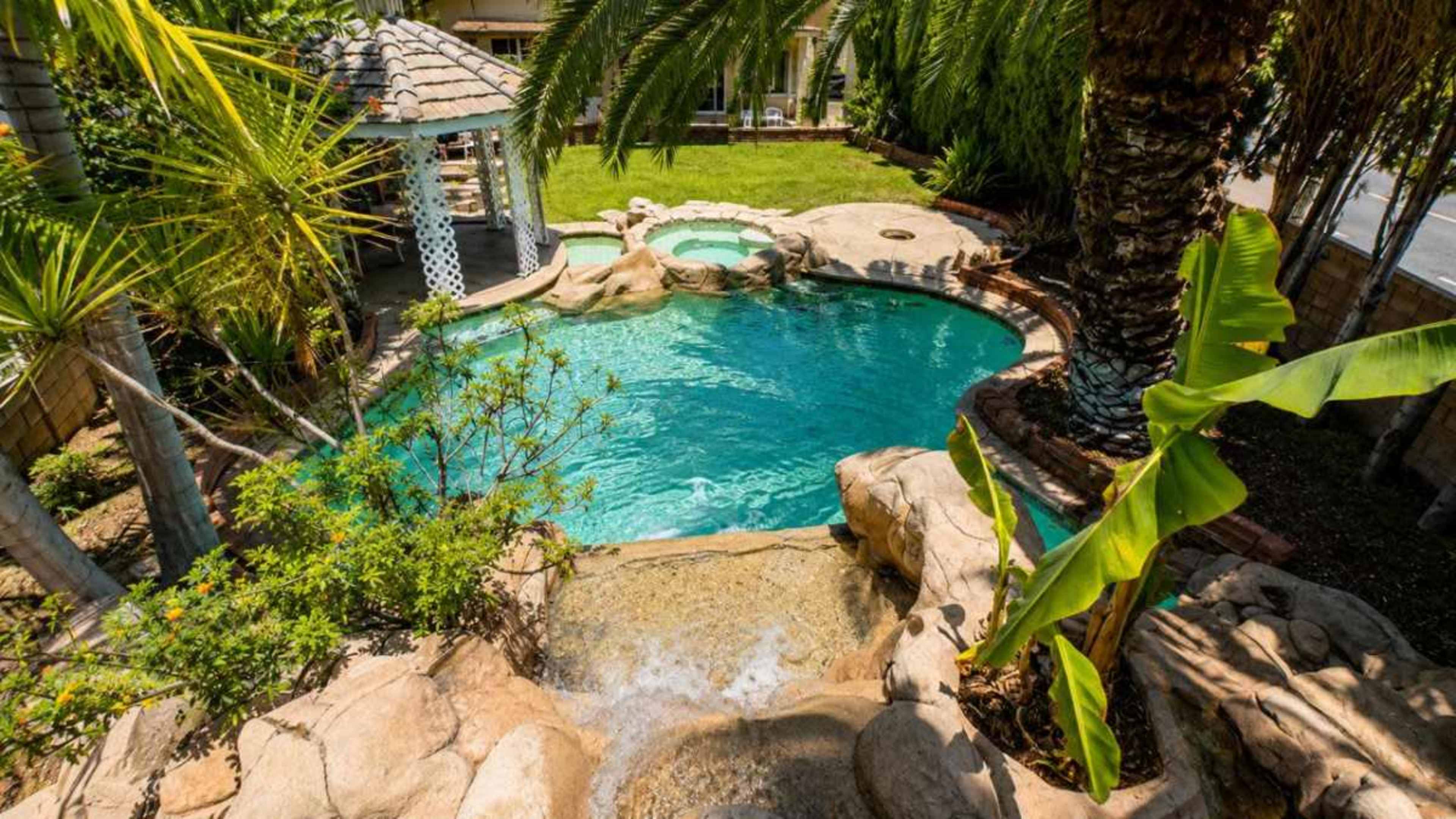 The image shows a backyard swimming pool with a waterfall feature, surrounded by palm trees and a grassy area.