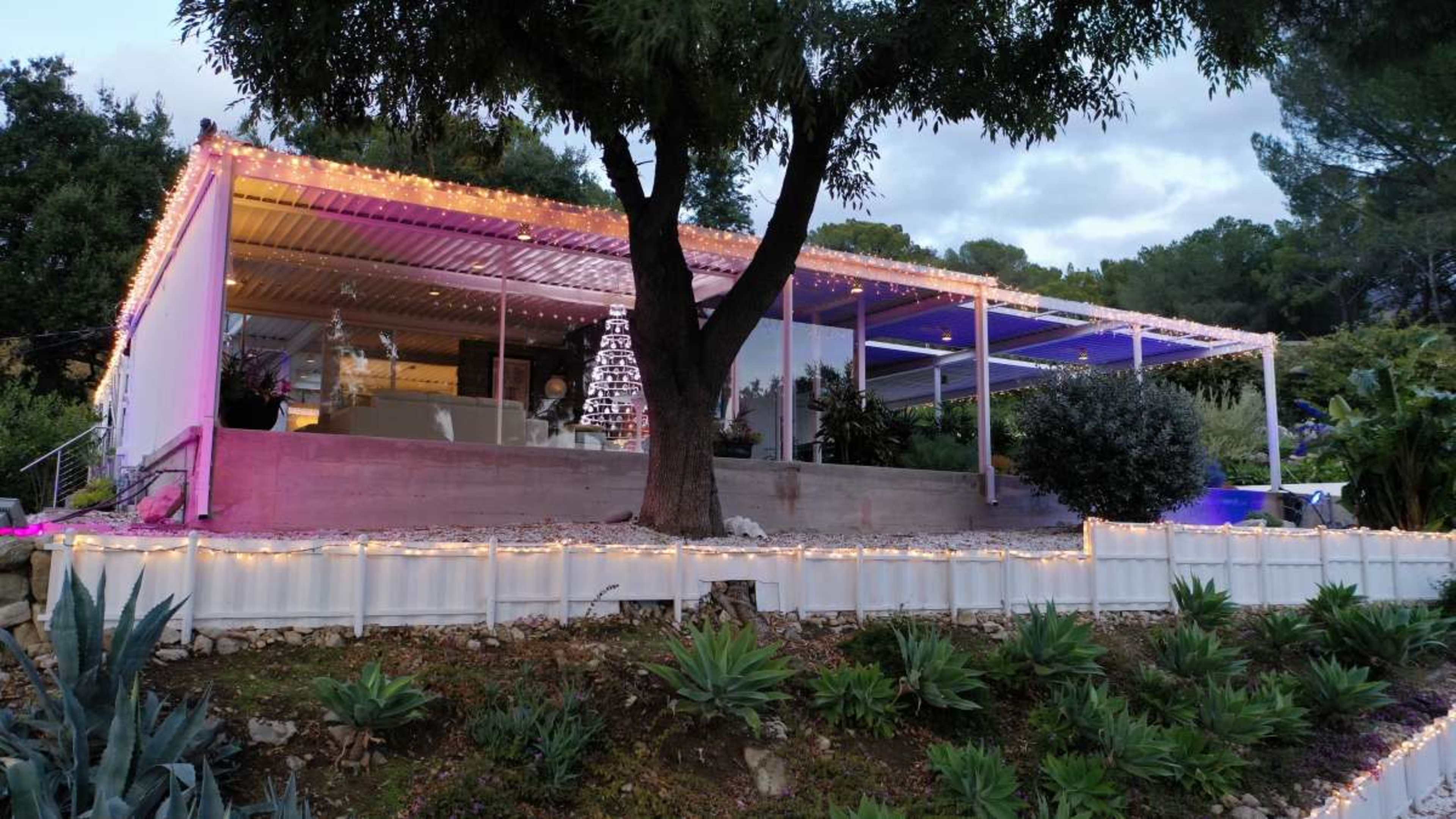 A modern house with large windows is surrounded by trees and landscaping, adorned with festive lights along its perimeter.