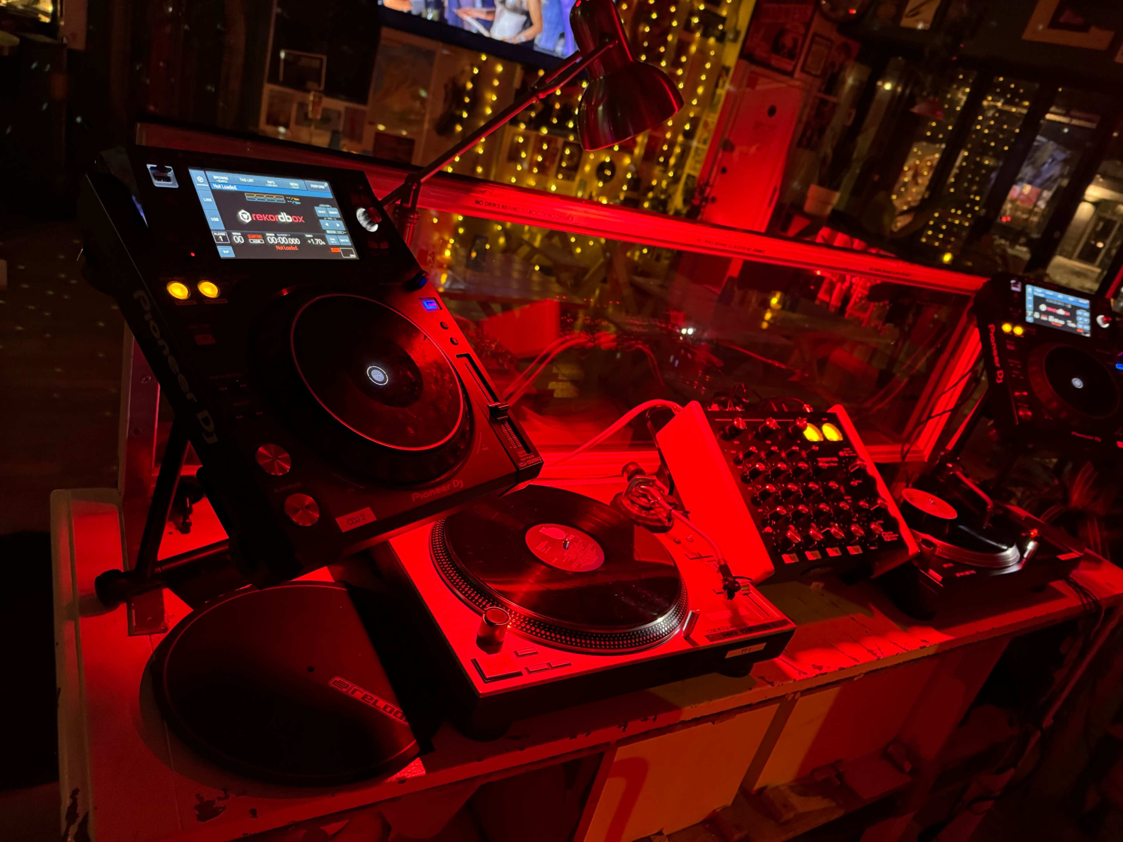 The image shows a DJ setup with turntables and a digital mixer illuminated by red lighting.