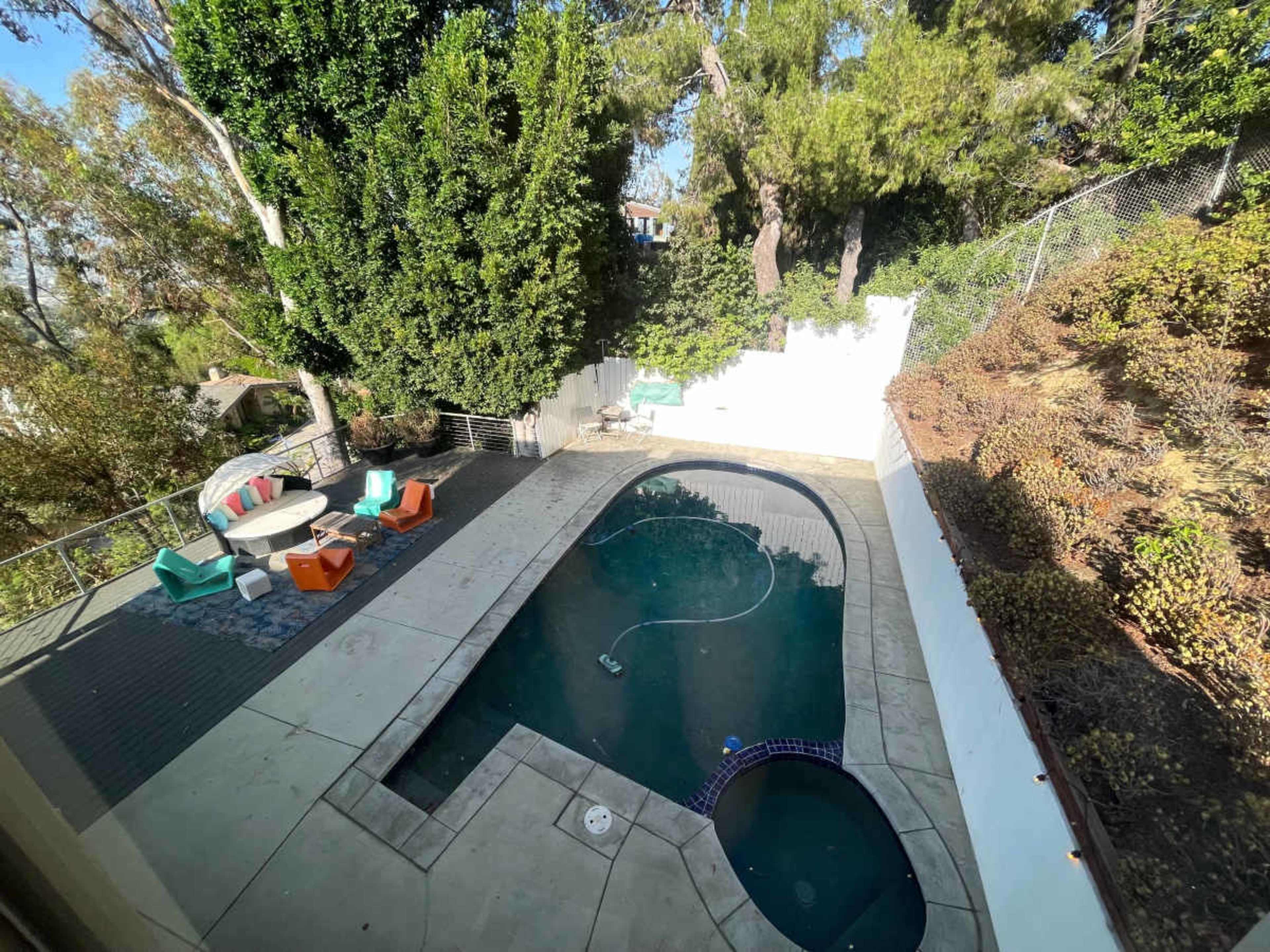 A backyard features a rectangular swimming pool surrounded by stone paving and colorful lounge chairs, with trees and shrubs providing greenery.