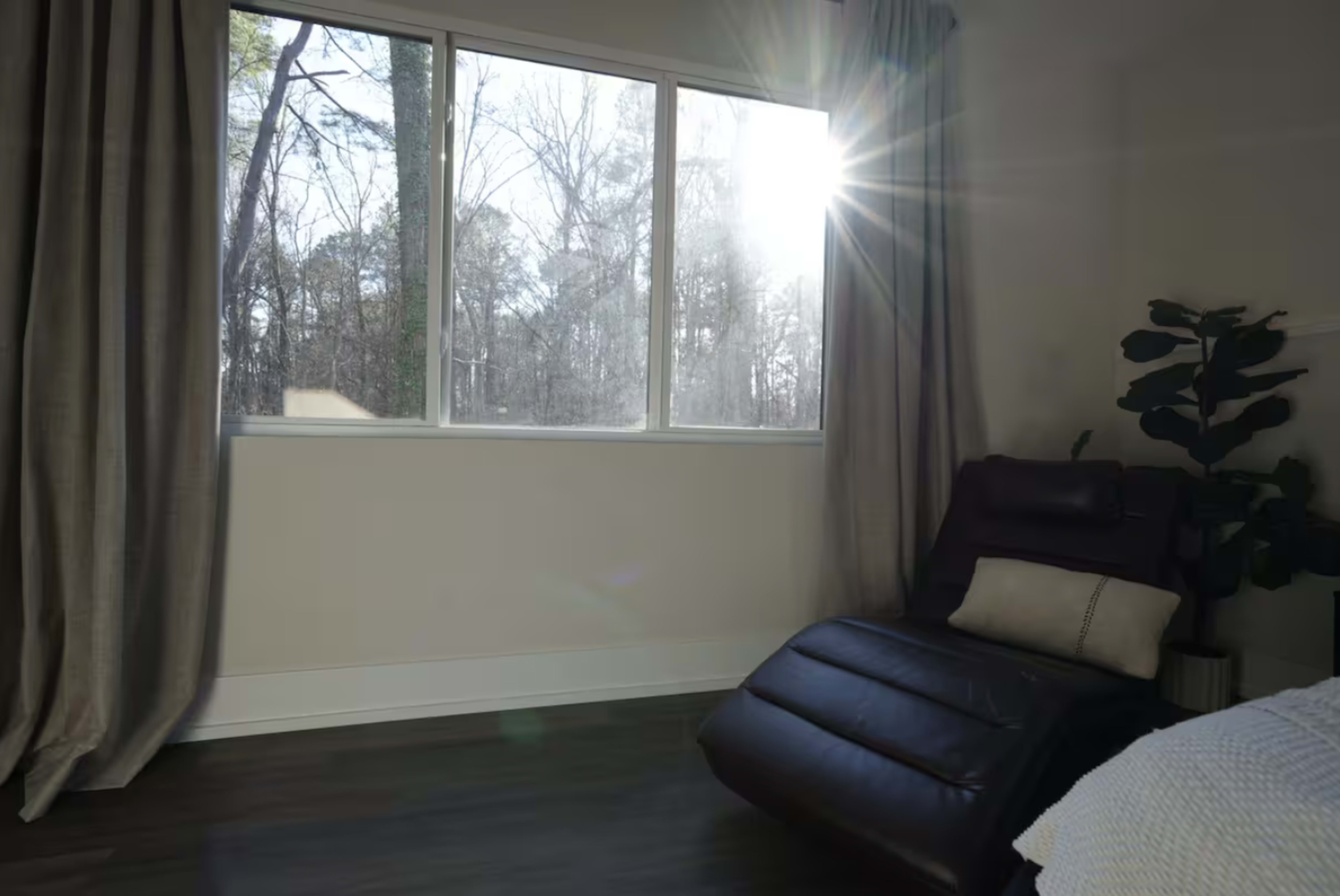A sunlit bedroom features a large window with sheer curtains, a reclining chair, and a potted plant.