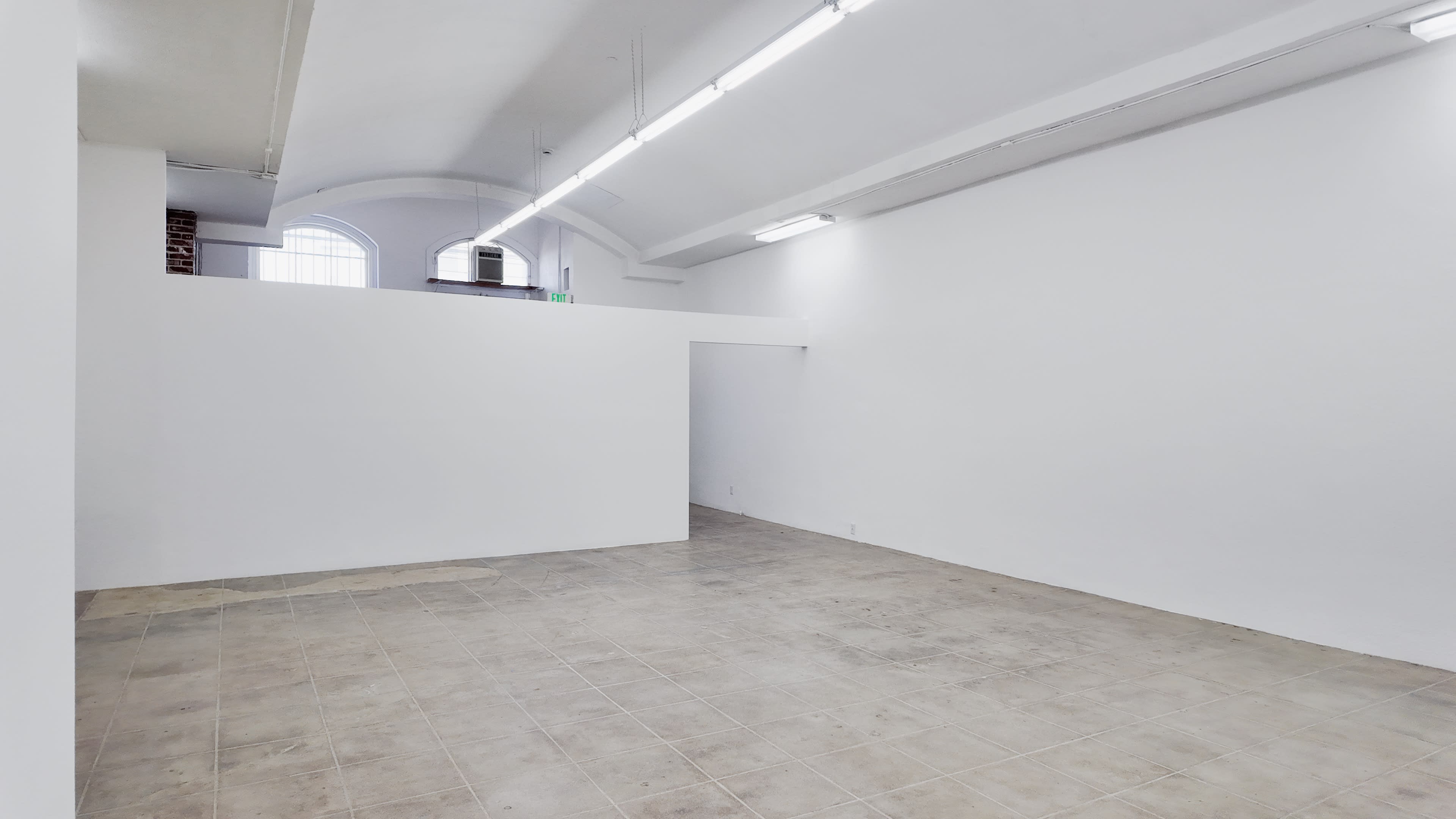 The image shows a spacious, empty room with white walls and a tiled floor, featuring overhead lighting and a doorway on one side.