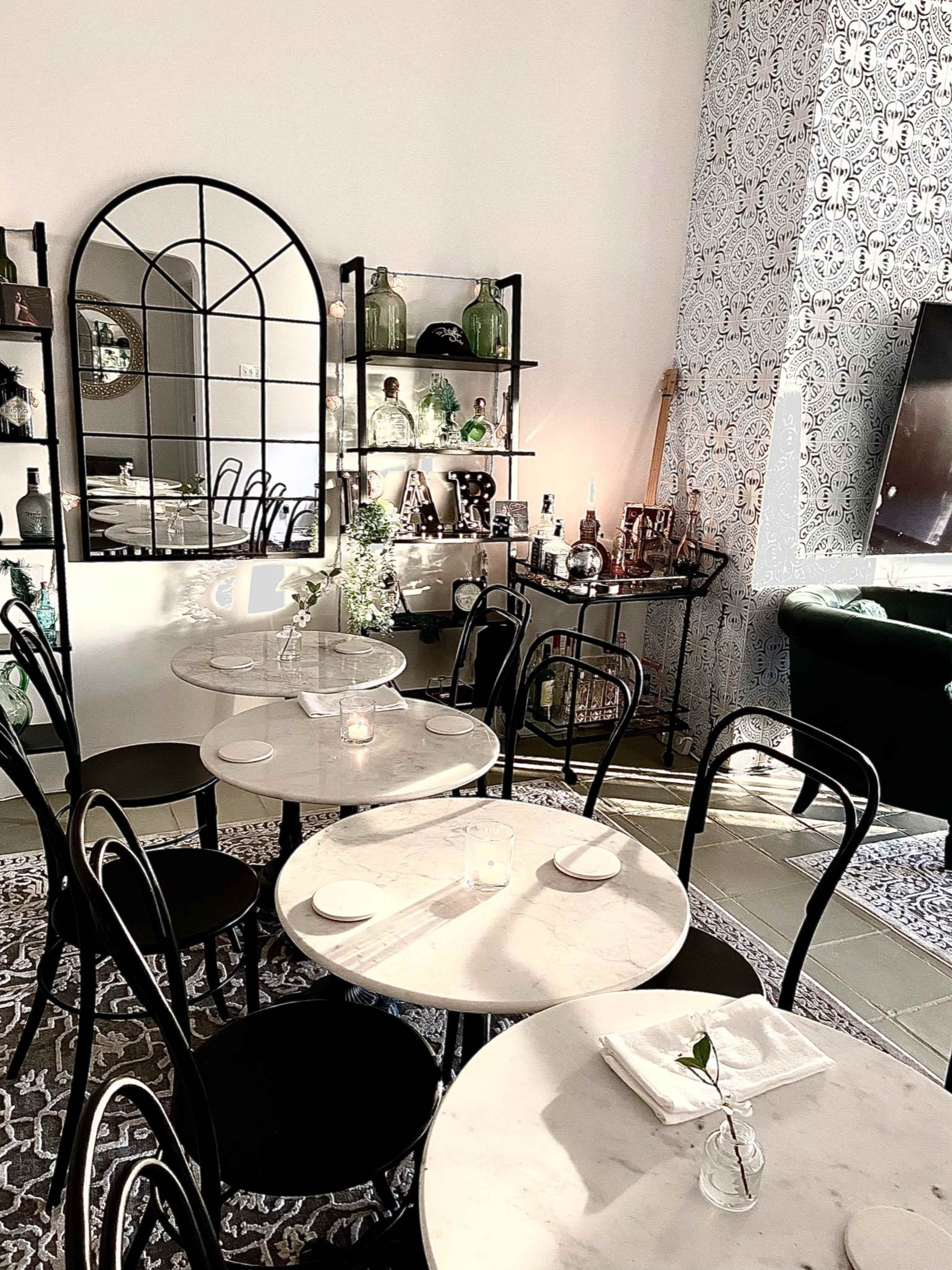 A bright café interior features several marble tables surrounded by black chairs, with a large mirror and shelves displaying various glassware and green decor.