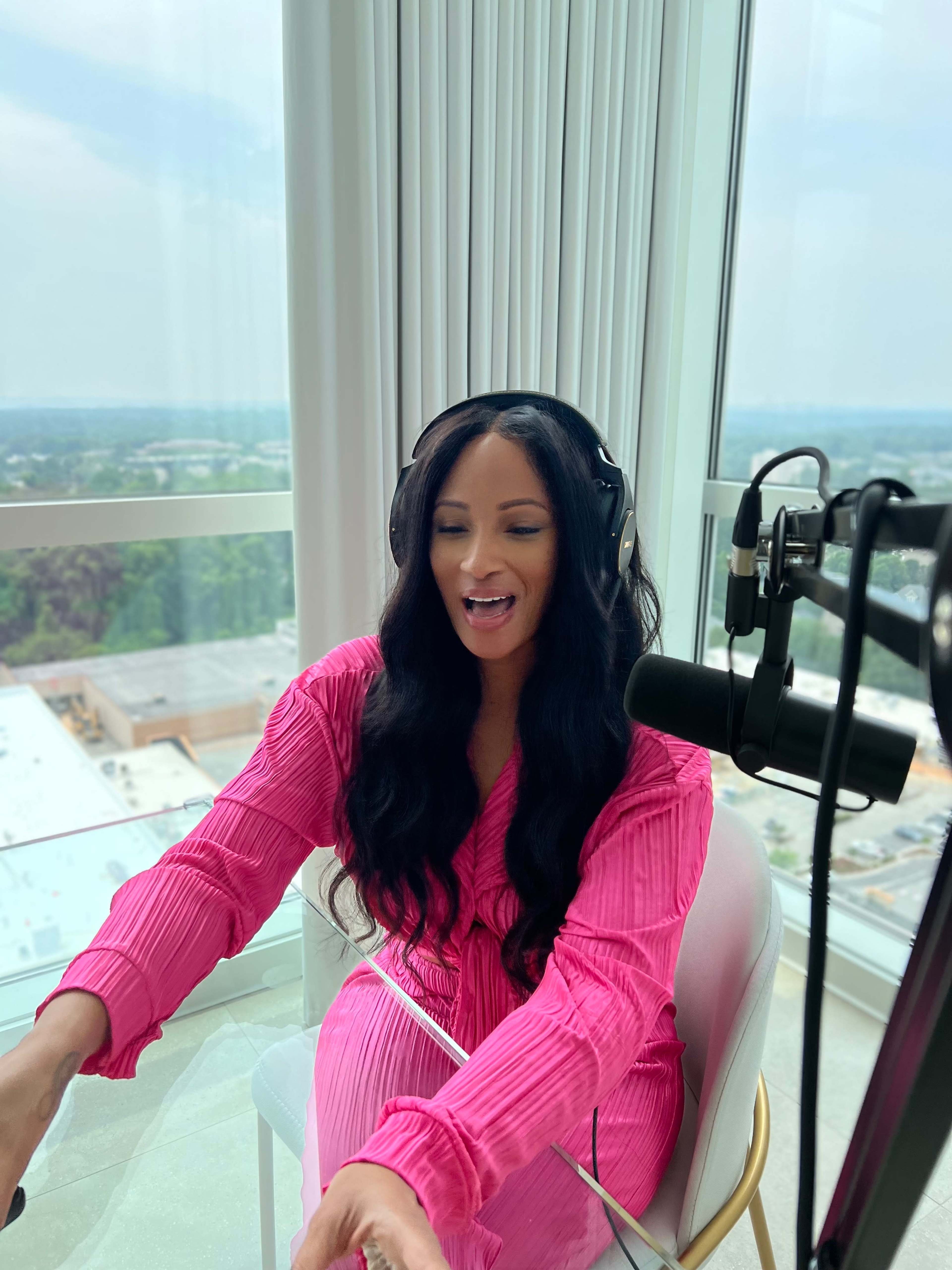 A woman in a pink dress is seated in front of a large window with a microphone and headphones, appearing to engage in a conversation or recording.