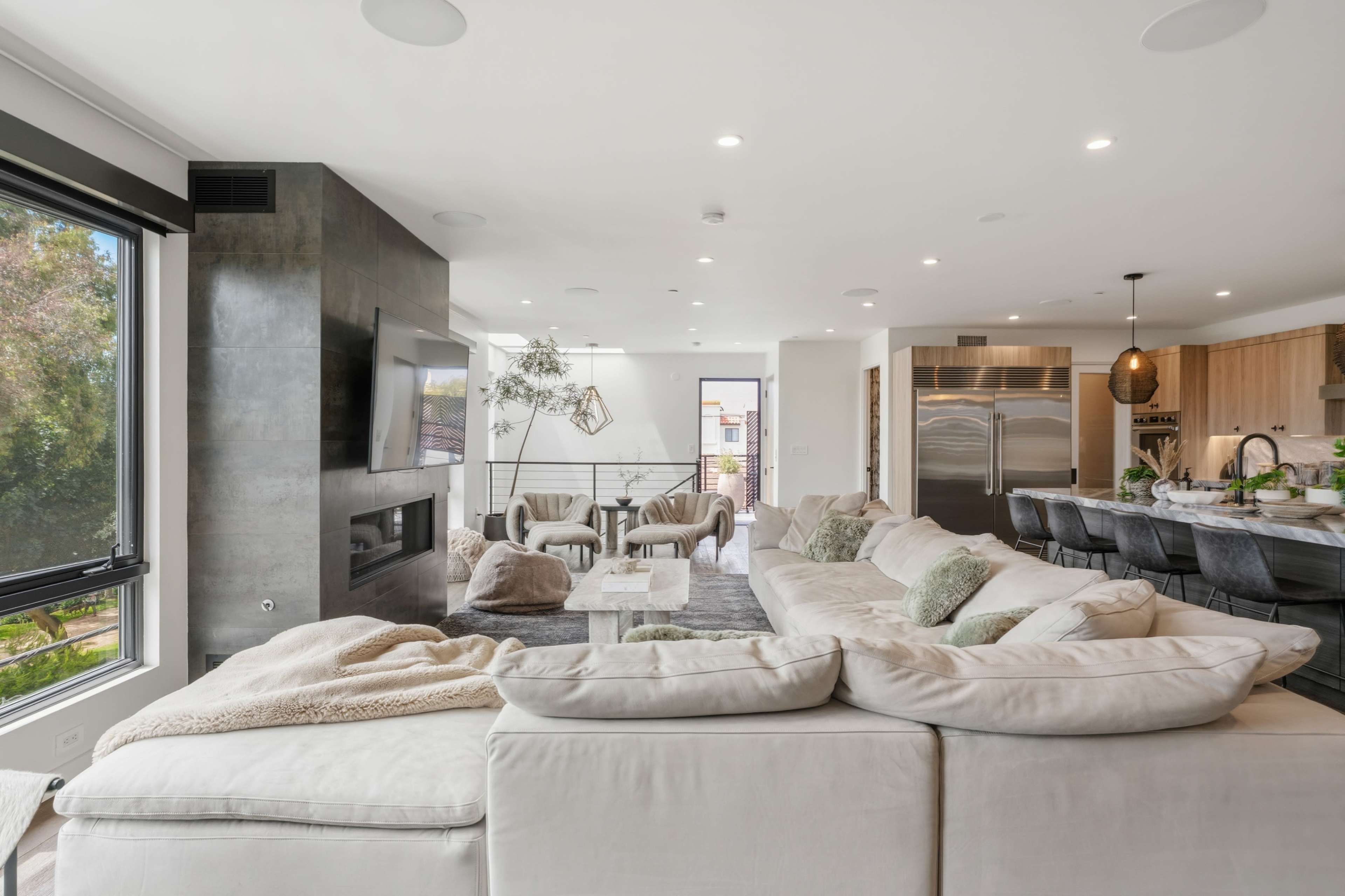 The image shows a modern living room with a large sectional sofa, a dining area, and an open kitchen, all featuring contemporary decor and natural light.