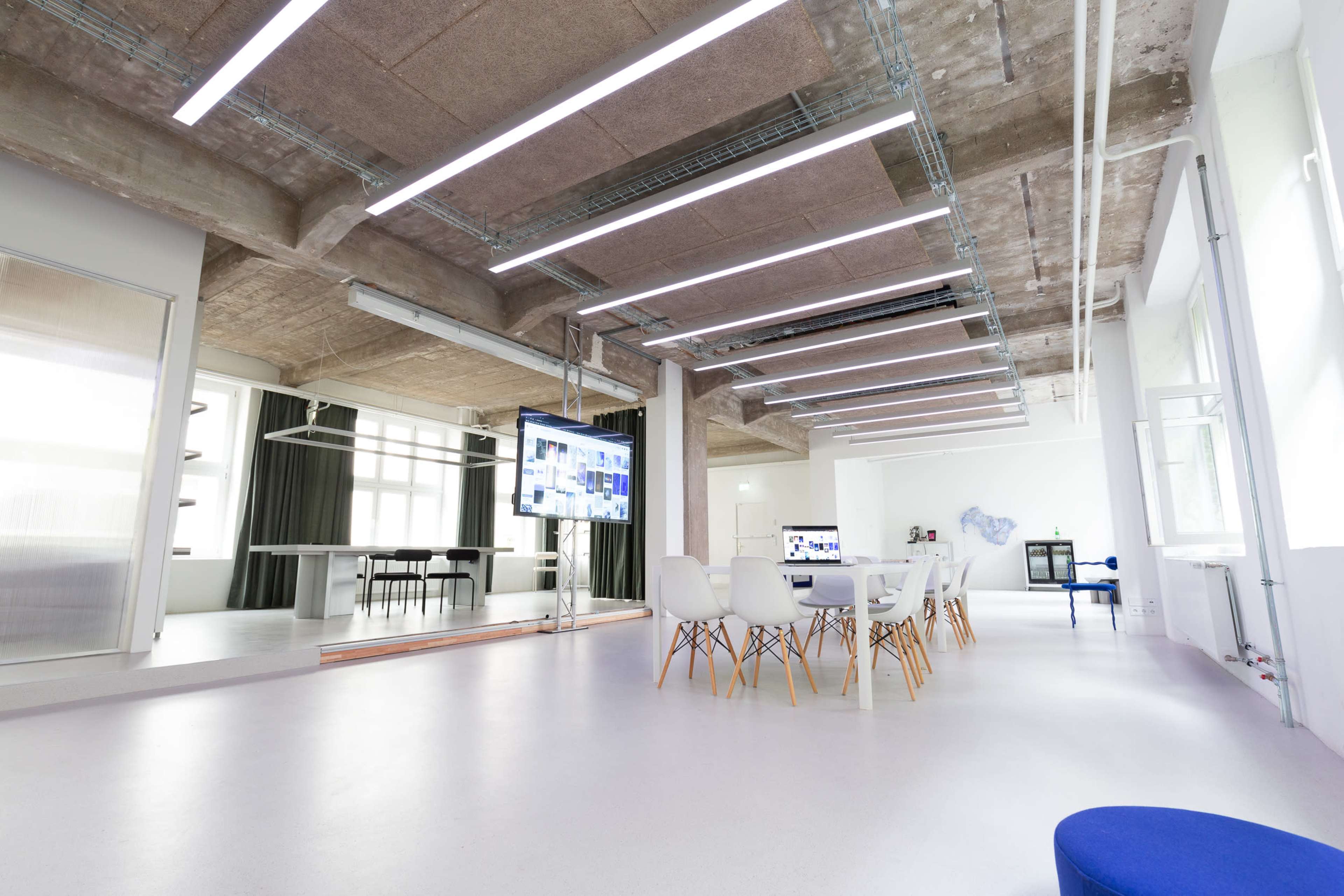 A modern office space with white walls, a large table surrounded by chairs, and a screen displaying digital content.