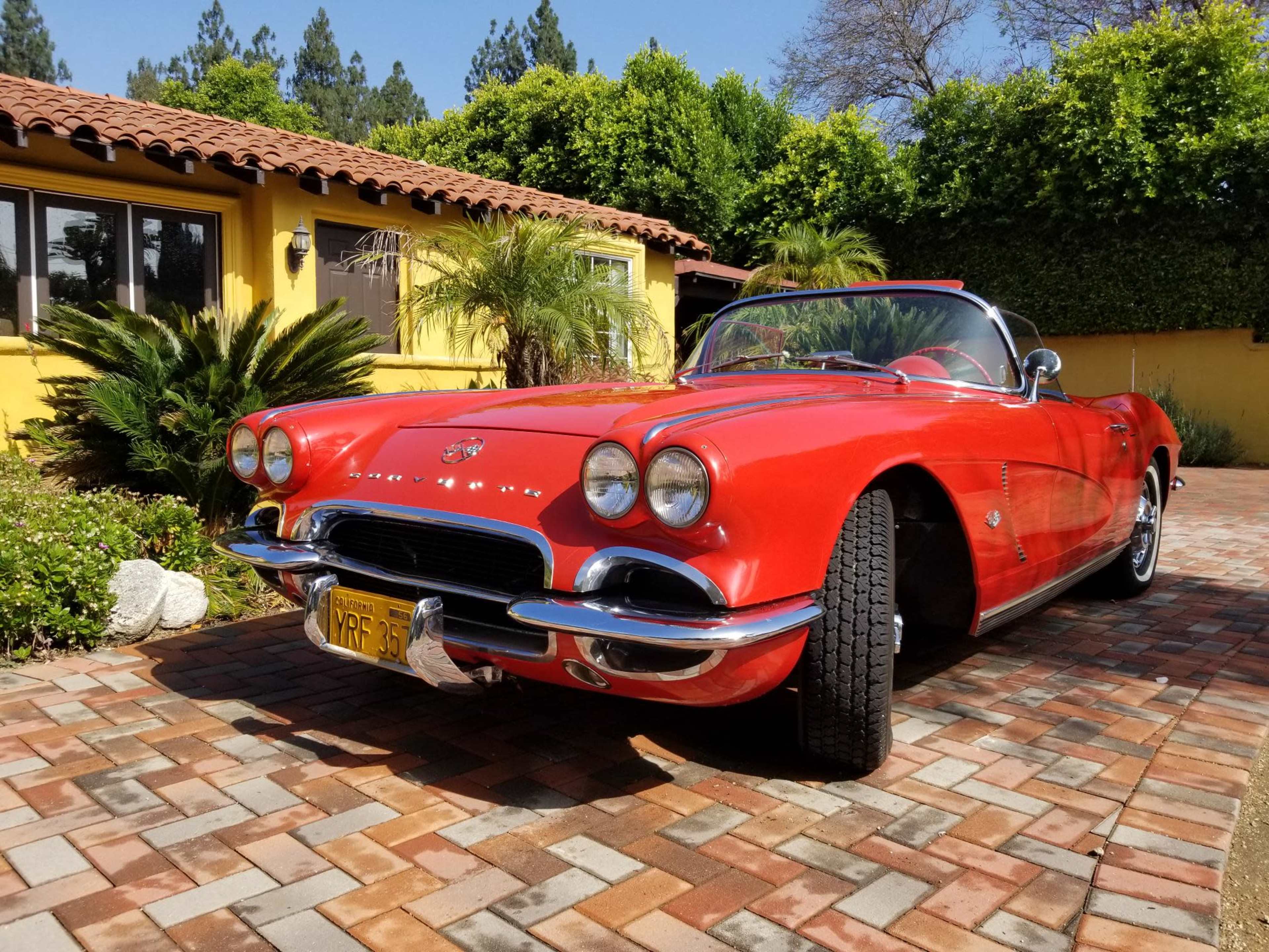 A red Chevrolet Corvette is parked on a brick driveway in front of a yellow house surrounded by greenery.
