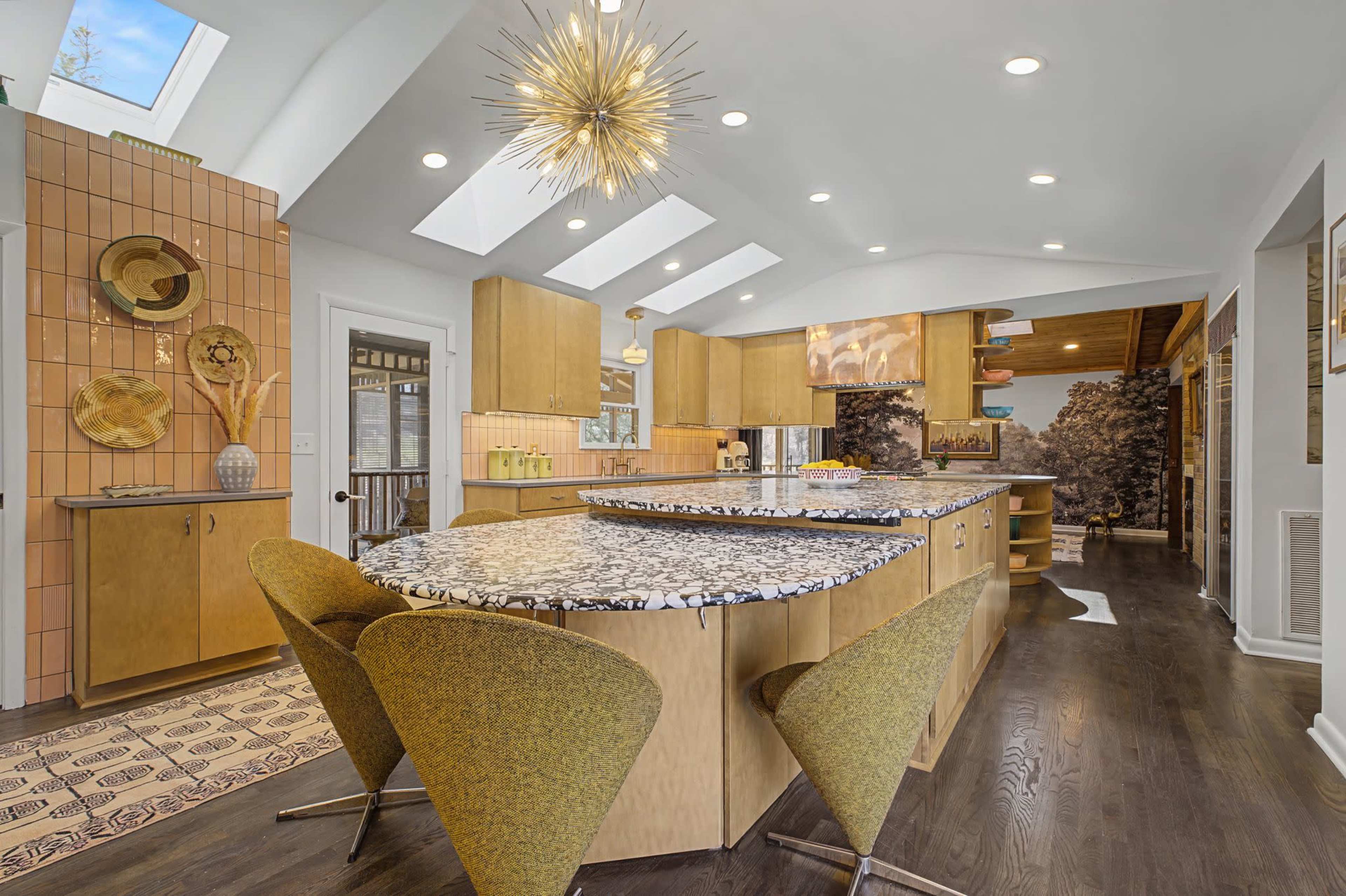 The image shows a modern kitchen with a large, curved island featuring a speckled countertop, surrounded by three green chairs, and equipped with wooden cabinets and skylights.