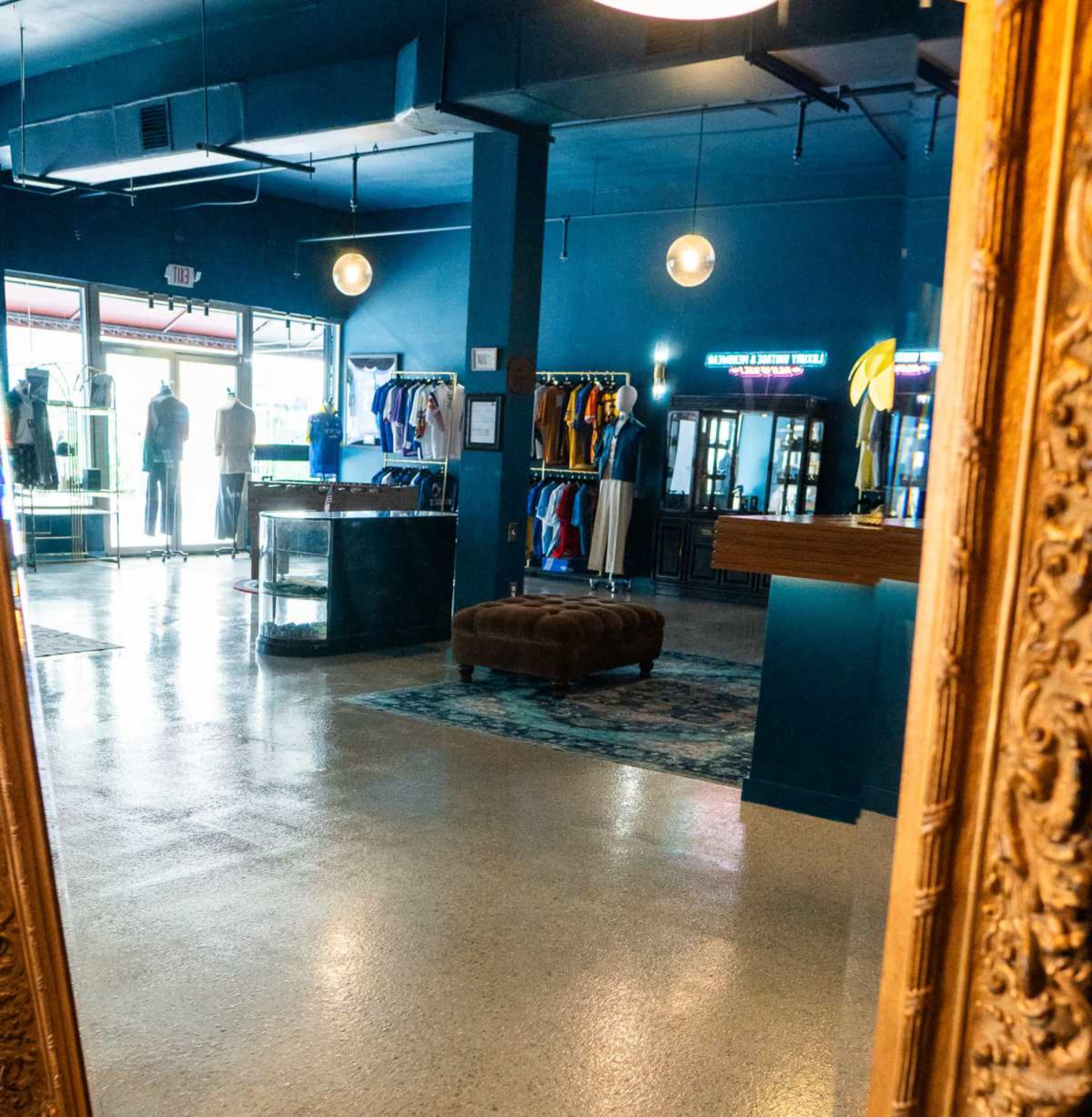 The image shows a clothing store interior with a blue wall, clothing racks, and mirrors, featuring a central seating area with a brown ottoman.