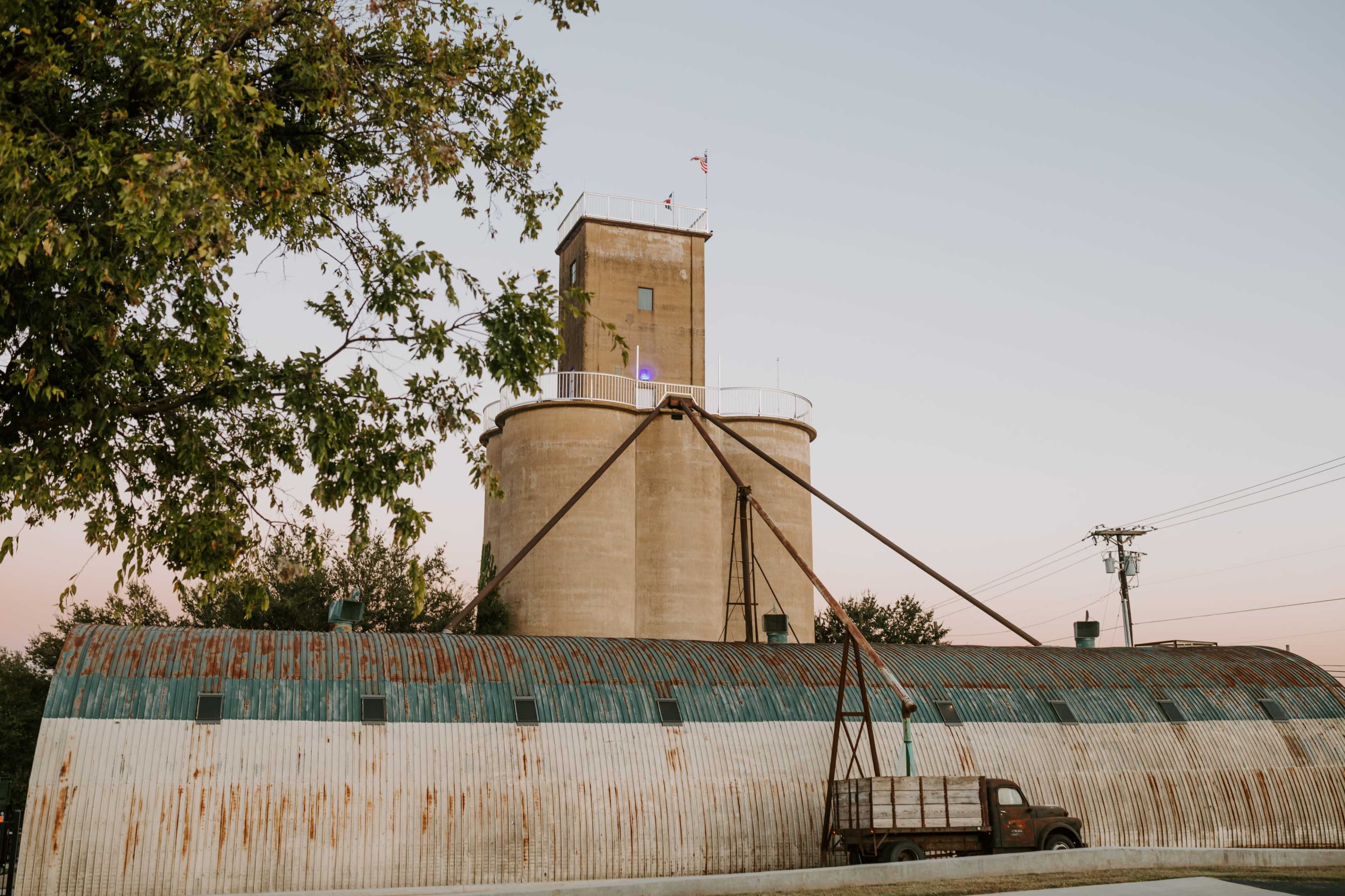 The image shows a large, cylindrical grain storage facility with a flat-roofed building and a vintage truck parked nearby.