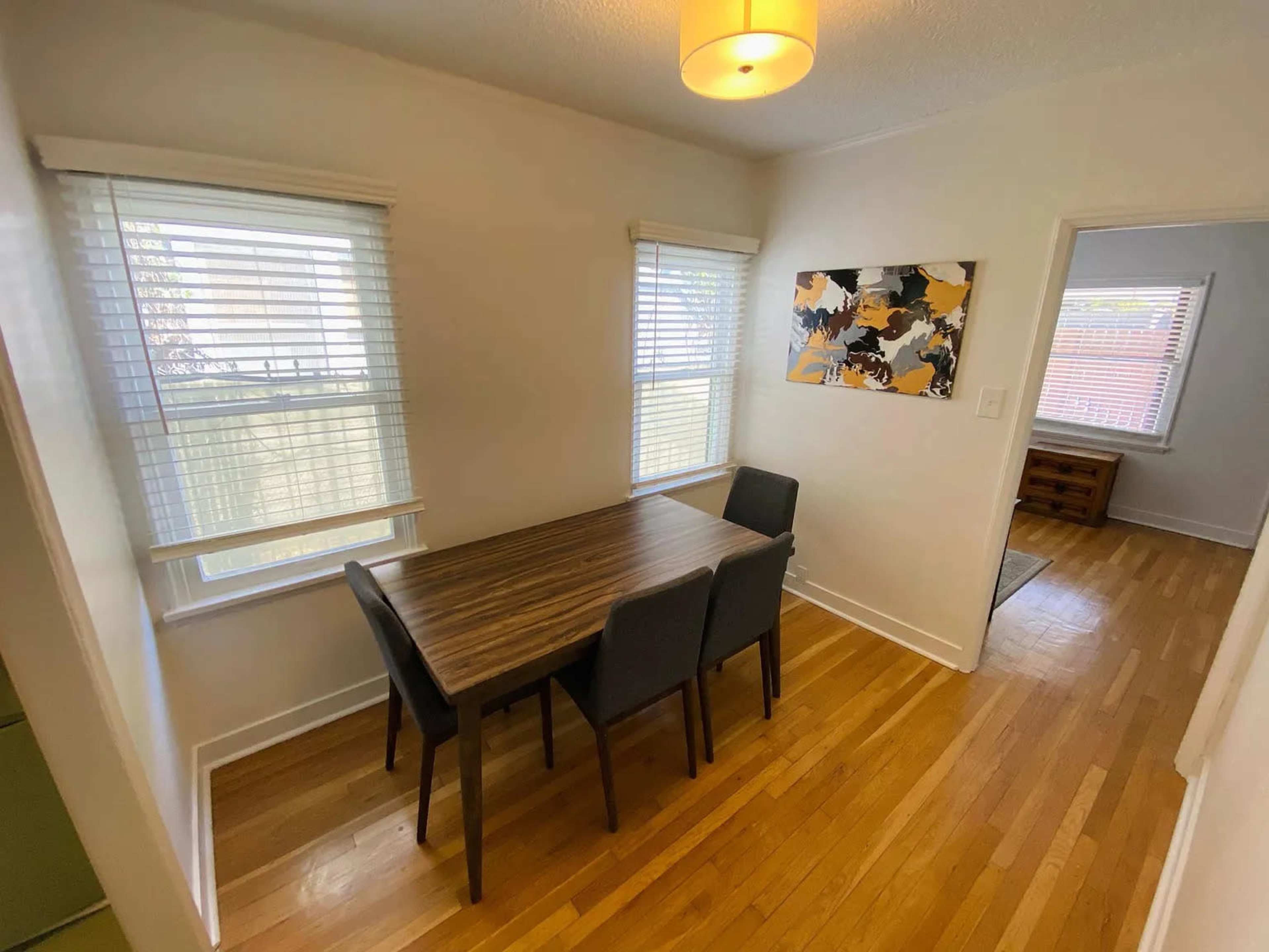 The image shows a dining area with a wooden table and four black chairs, illuminated by a ceiling light, alongside two windows with white blinds and an abstract painting on the wall.