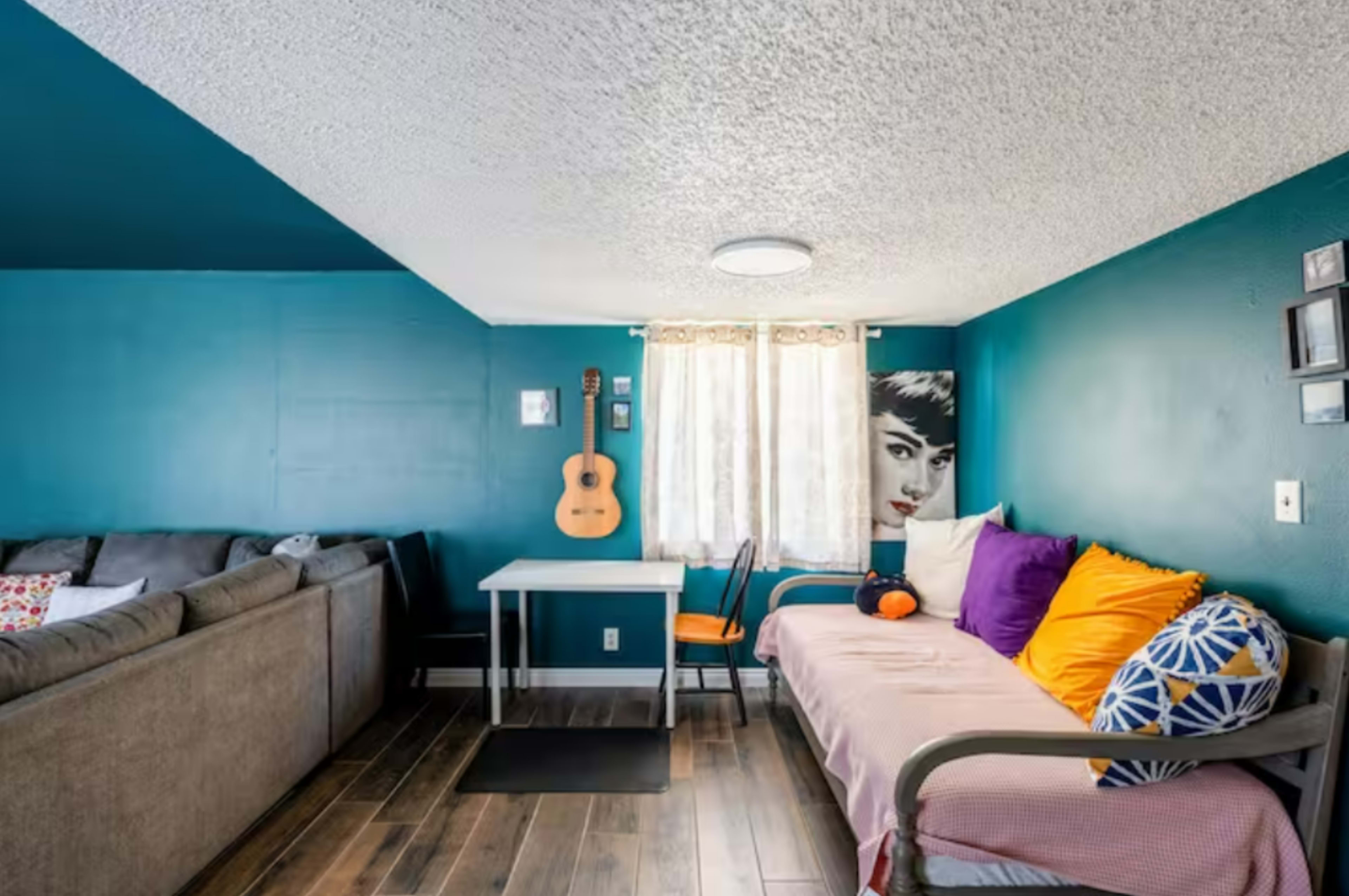 A small living space featuring a light teal wall, a couch with colorful pillows, a table with chairs, and a guitar hanging on the wall next to a window with sheer curtains.