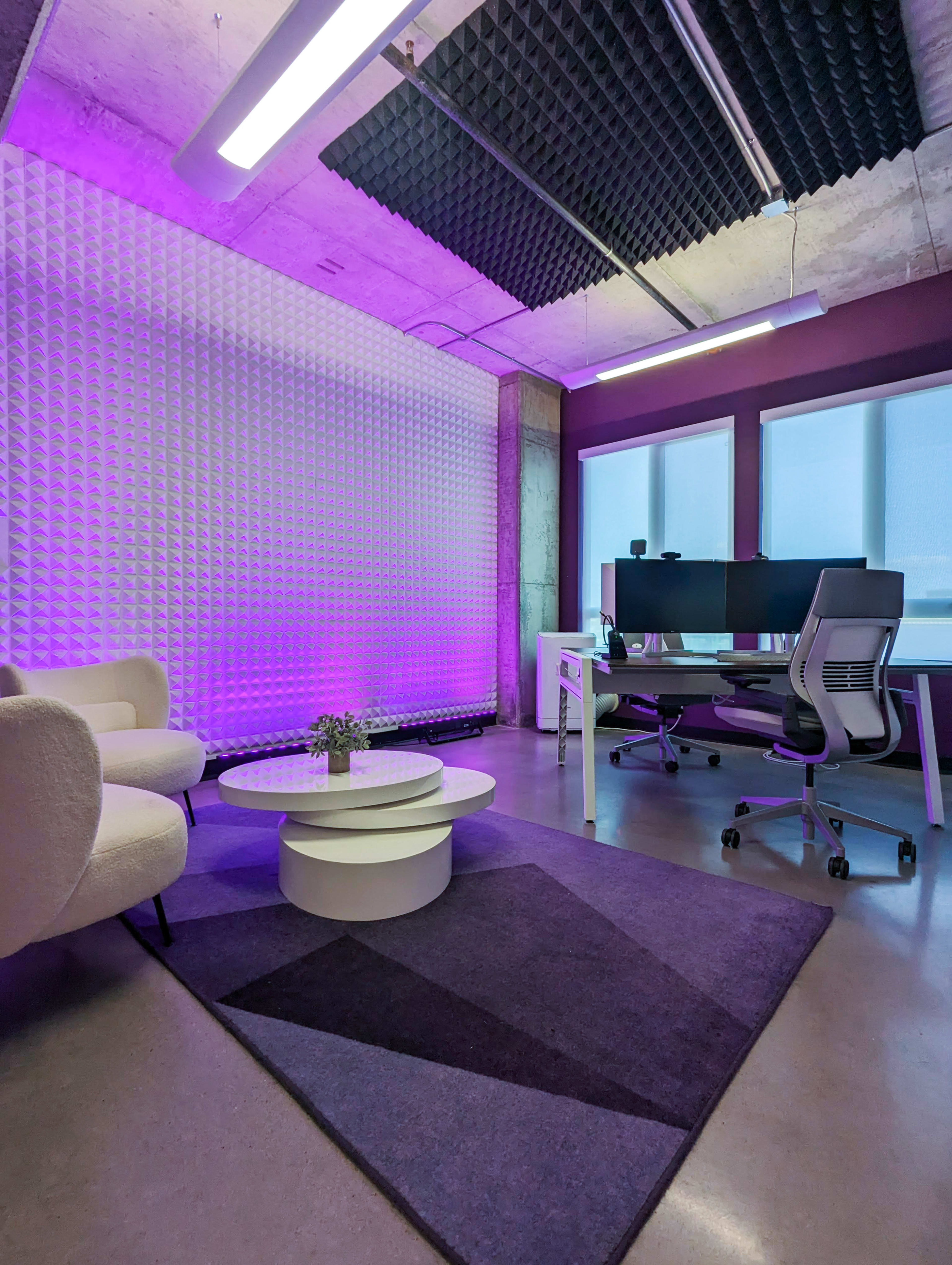 The image shows a modern office space featuring a seating area with a circular coffee table, a large desk with dual monitors, and purple accent lighting along the walls.