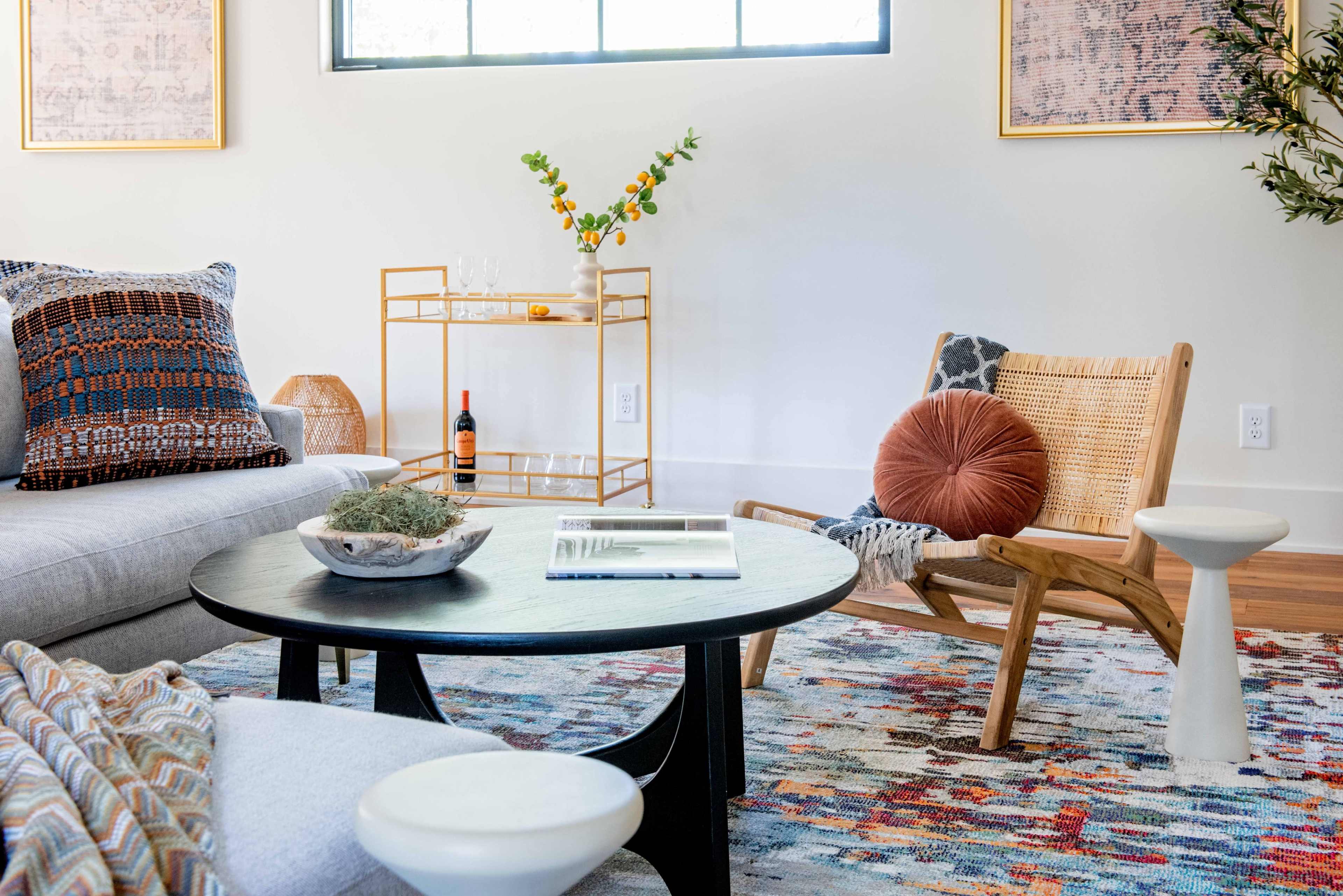 The image shows a modern living room featuring a gray sofa, a round black coffee table, a wooden chair with a cushion, and a decorative cart with a bottle of wine and a small vase.