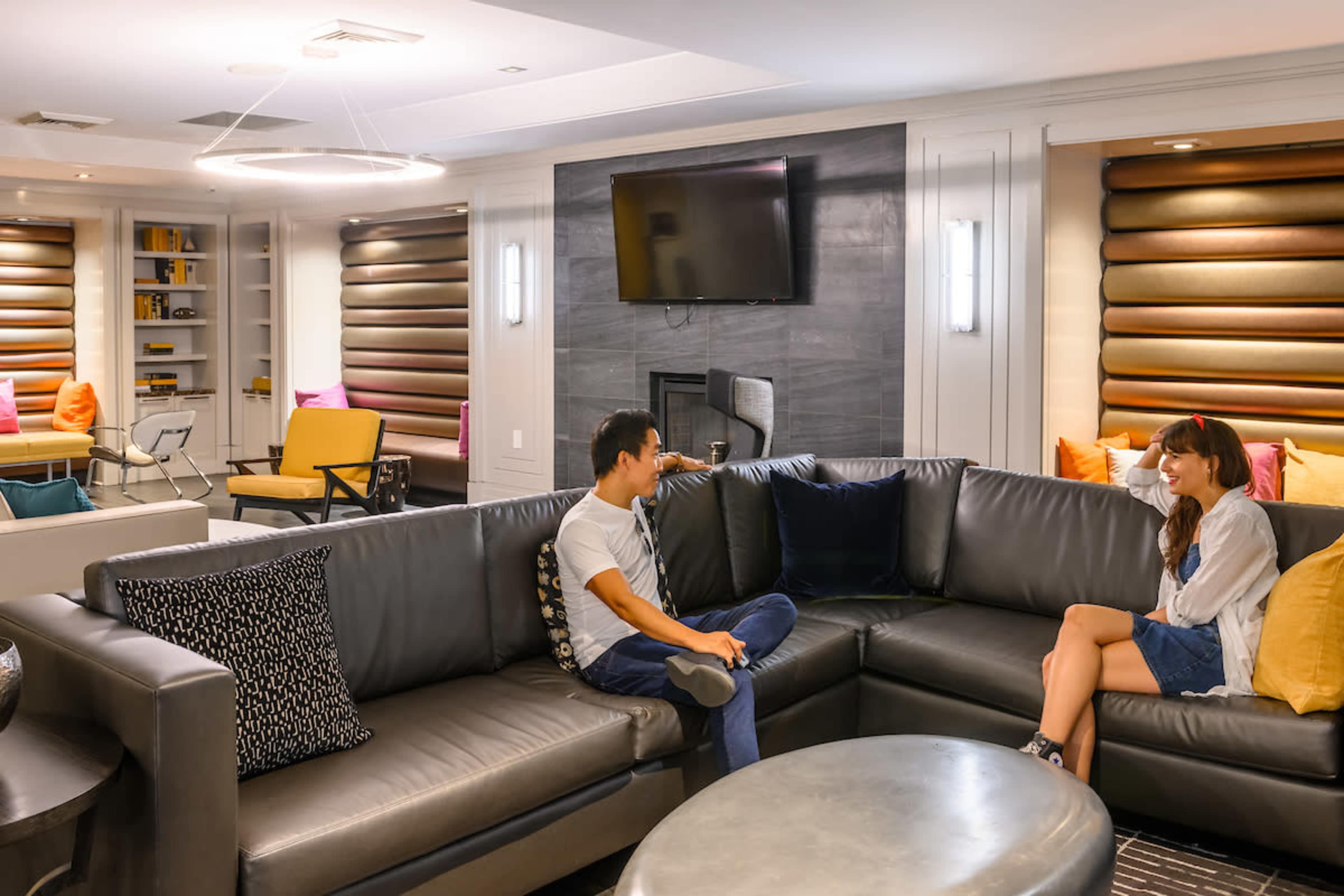 A man and a woman are seated on a sectional sofa in a modern lounge area with a large television mounted on the wall.