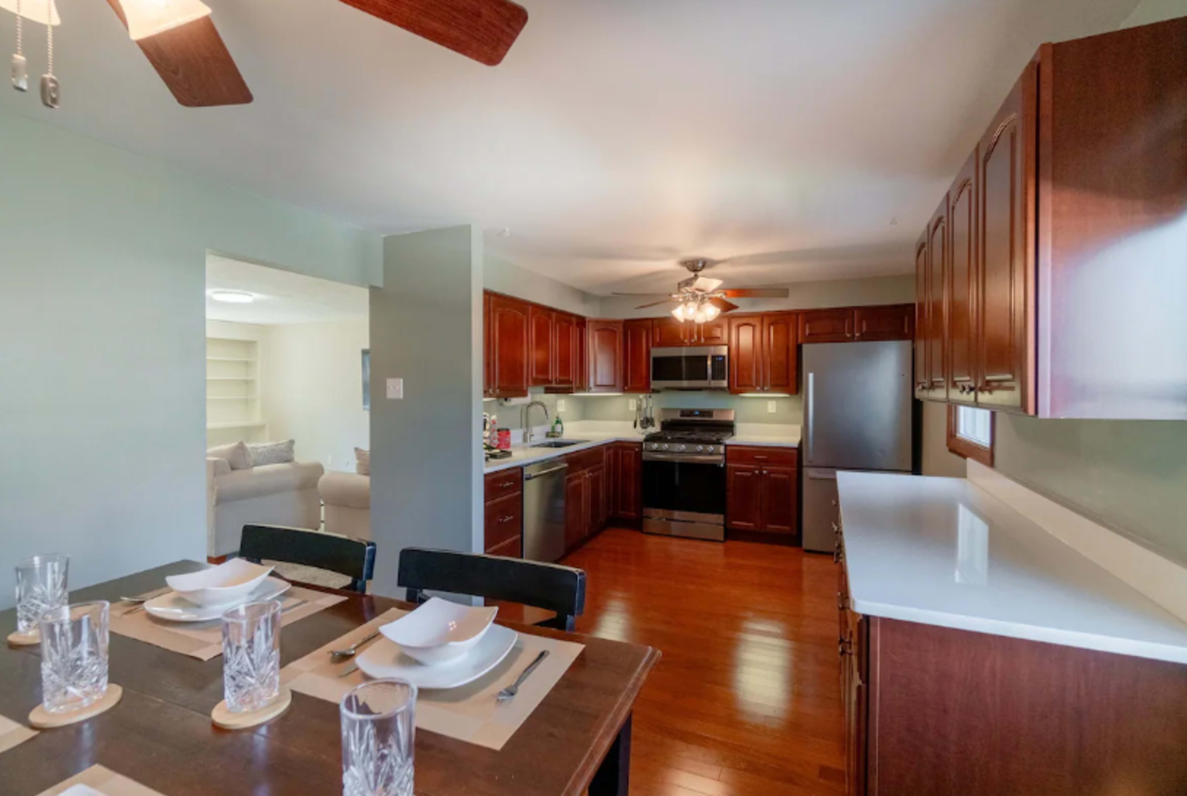 The image shows a modern kitchen with dark wood cabinetry, stainless steel appliances, and a dining area featuring a wooden table set with dishware.