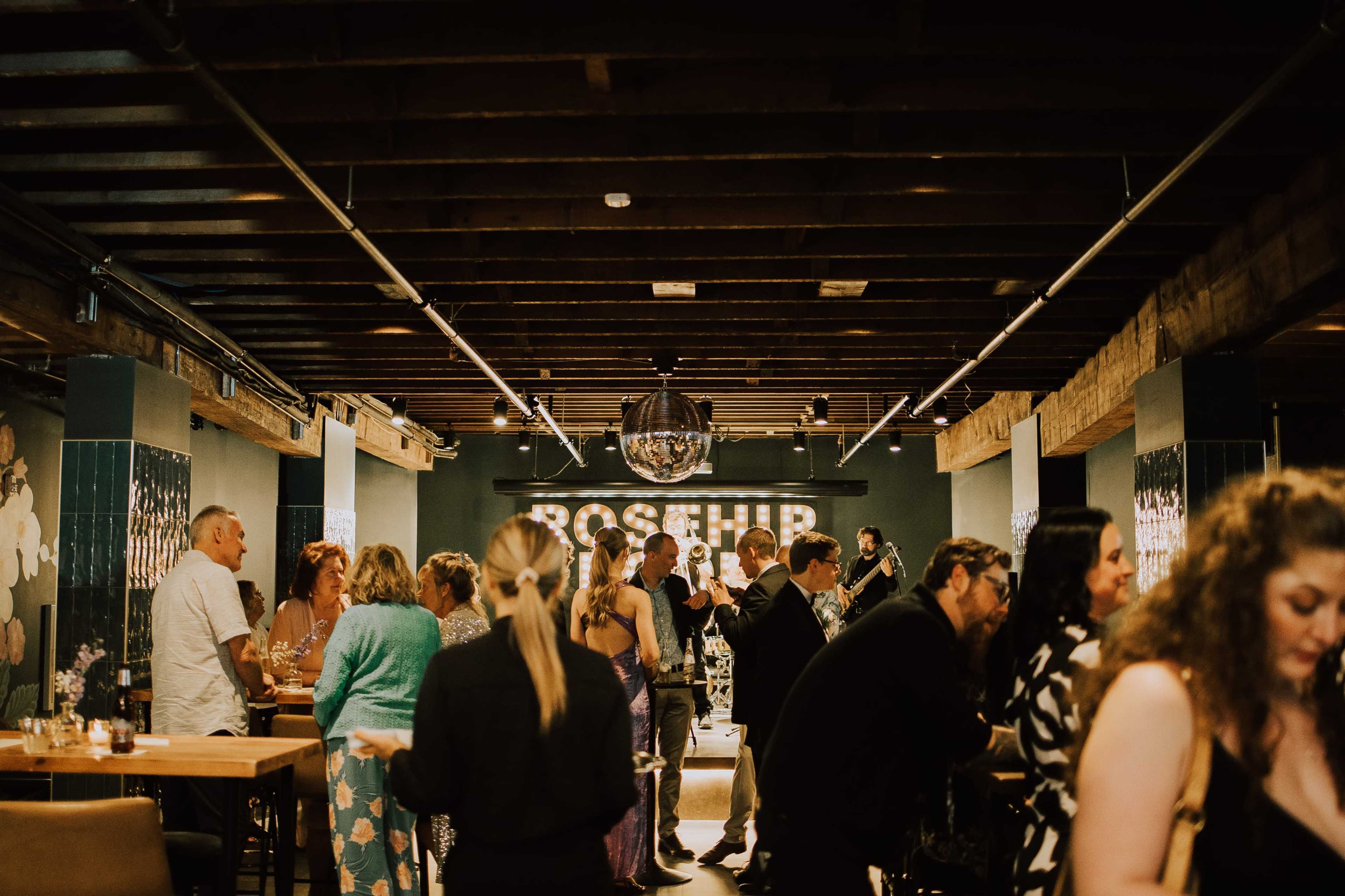 A lively gathering takes place in a stylish bar with guests mingling at tables and a band performing in the background.