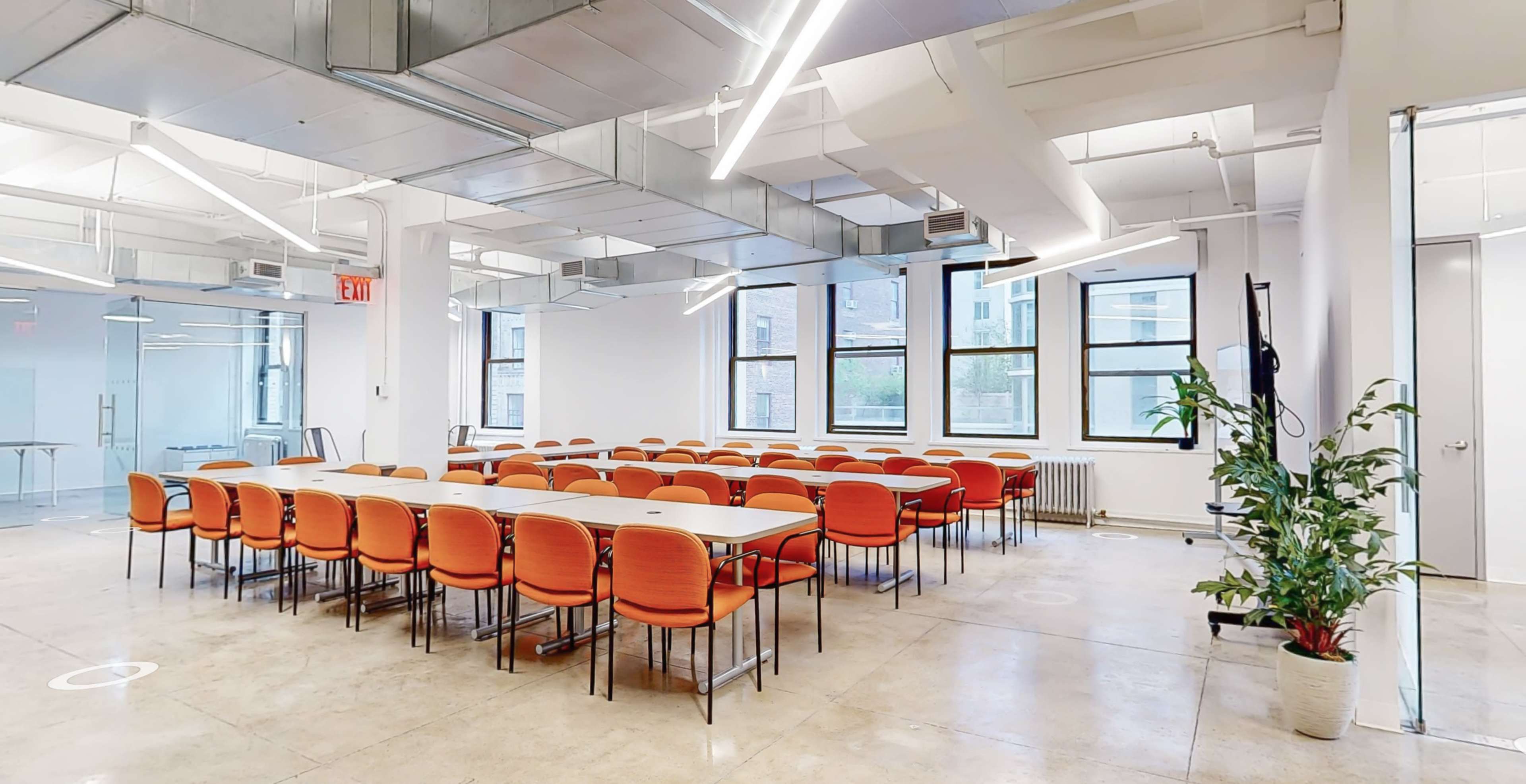 A modern conference room with a long table surrounded by orange chairs, large windows, and bright overhead lighting.