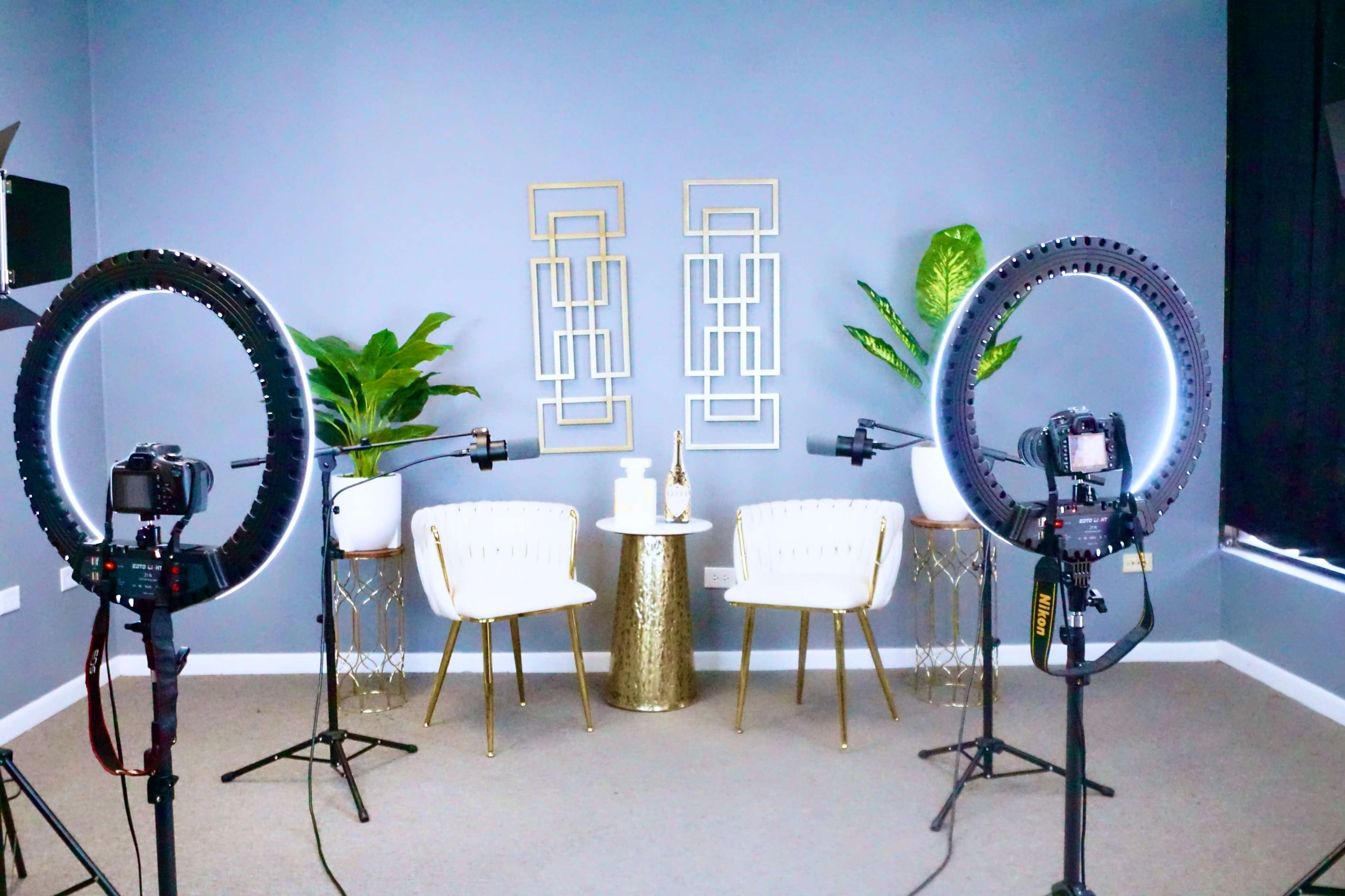The image shows a well-lit studio setup with two chairs, a small table, and decorative plants, surrounded by camera equipment and ring lights.