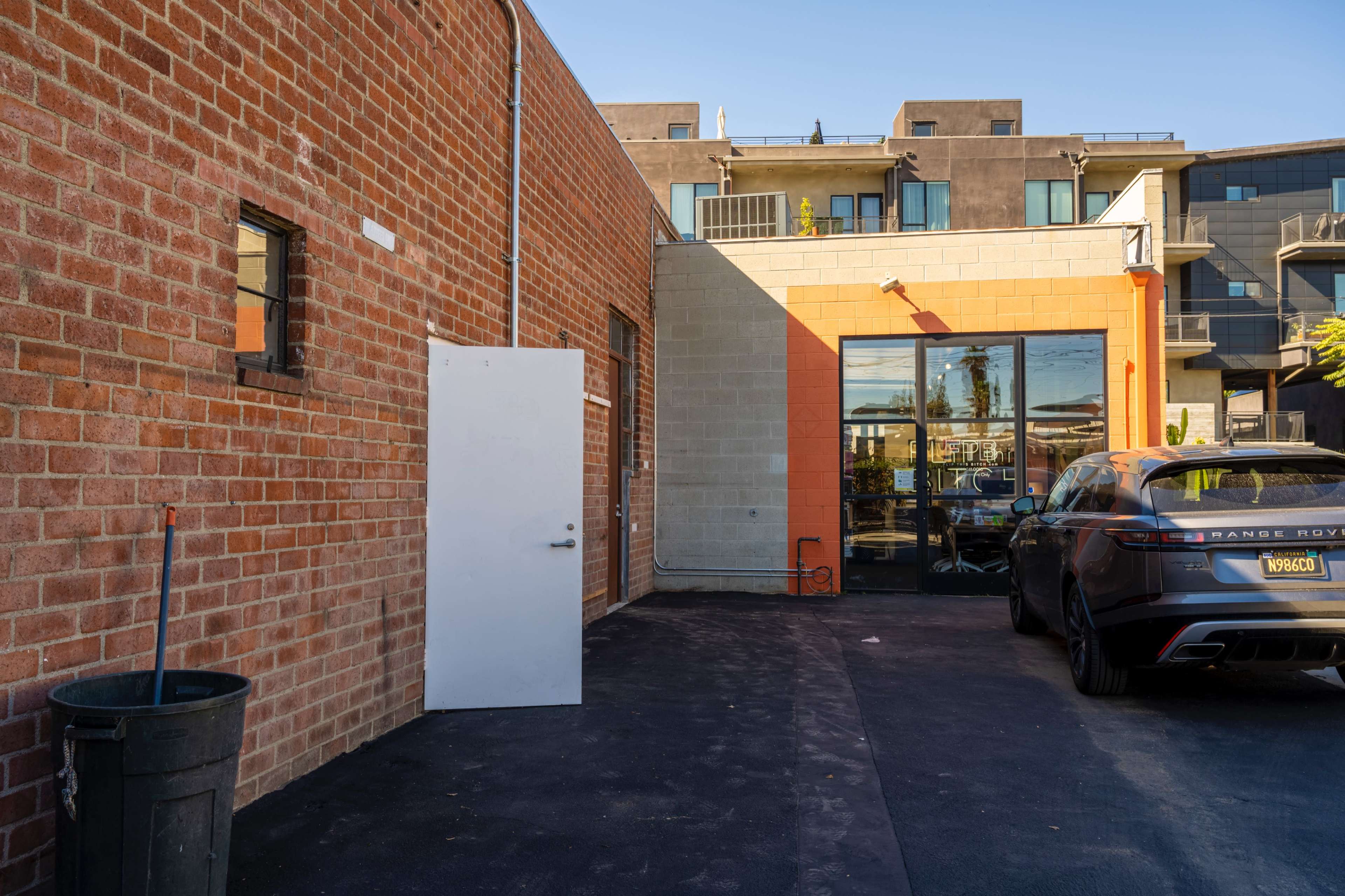 The image shows a brick wall with a white door beside a modern building and a parked Range Rover on a black asphalt surface.