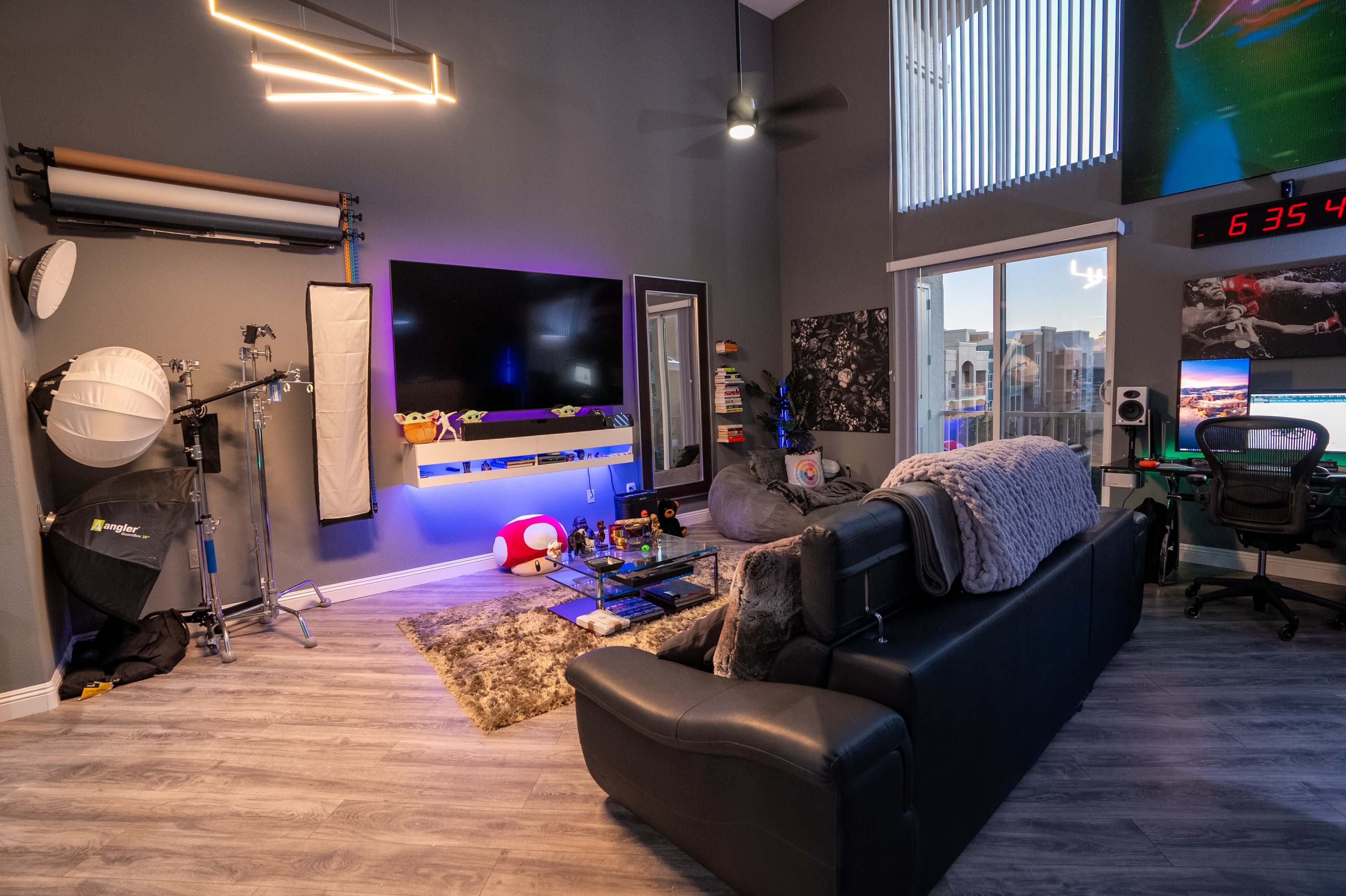 A modern living space featuring a black leather sofa, a wall-mounted television with colorful lighting, various decor items, and a workspace with a computer setup.