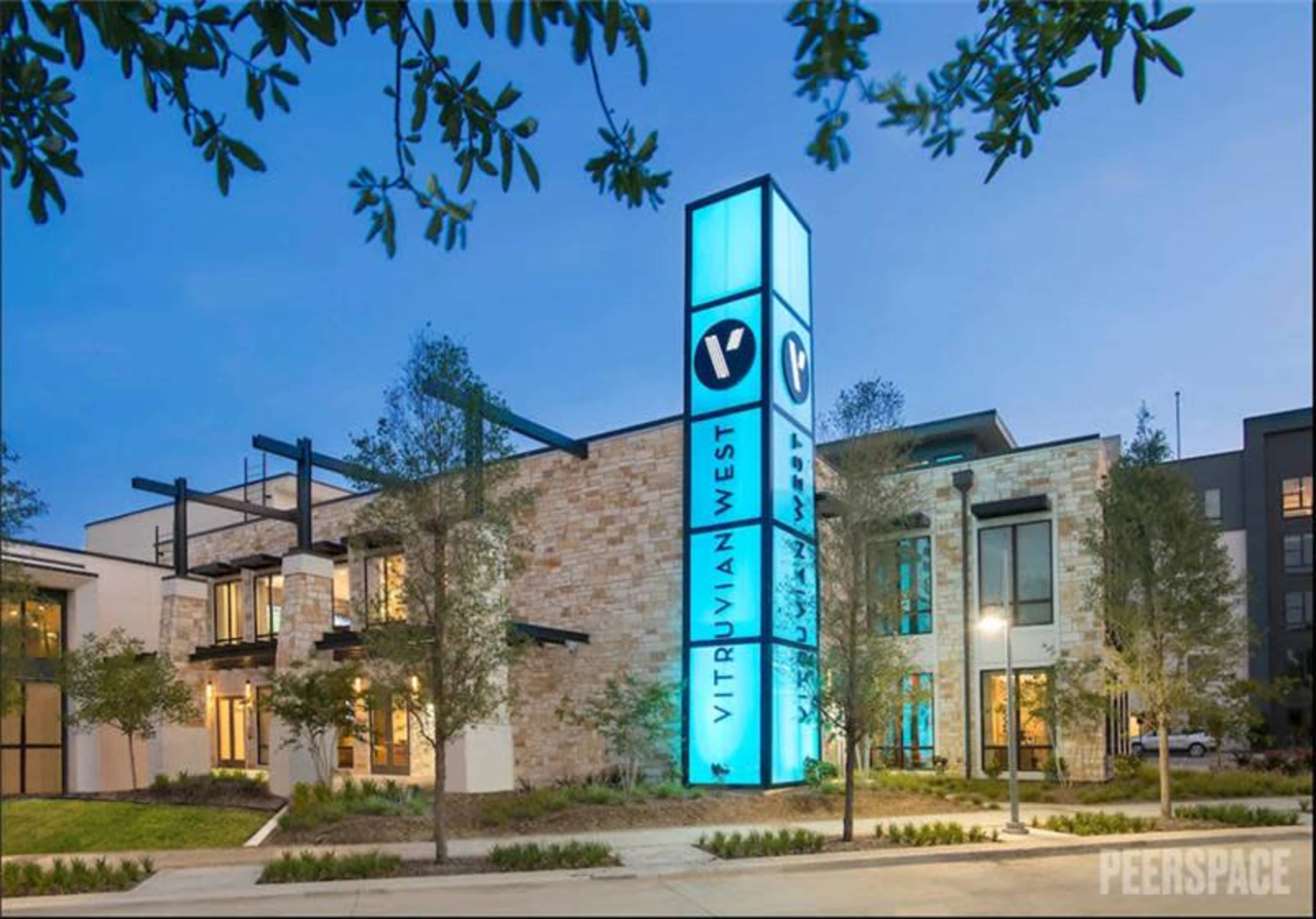 The image shows a modern building named "Vitruvian West," featuring a large illuminated sign, landscaping with small trees, and large windows.