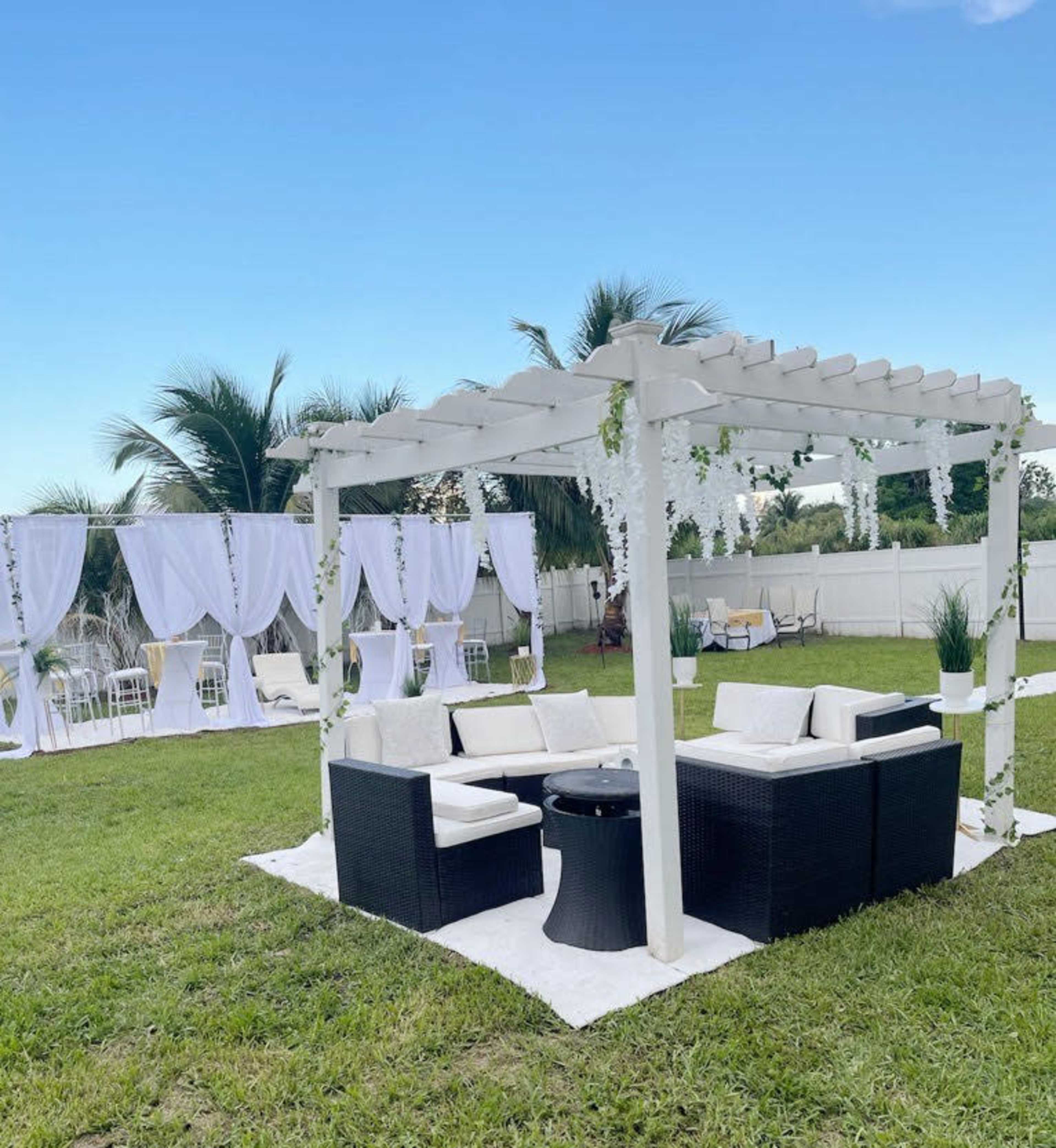 A white pergola with seating arranged on a grassy area, surrounded by white draped decorations and palm trees.