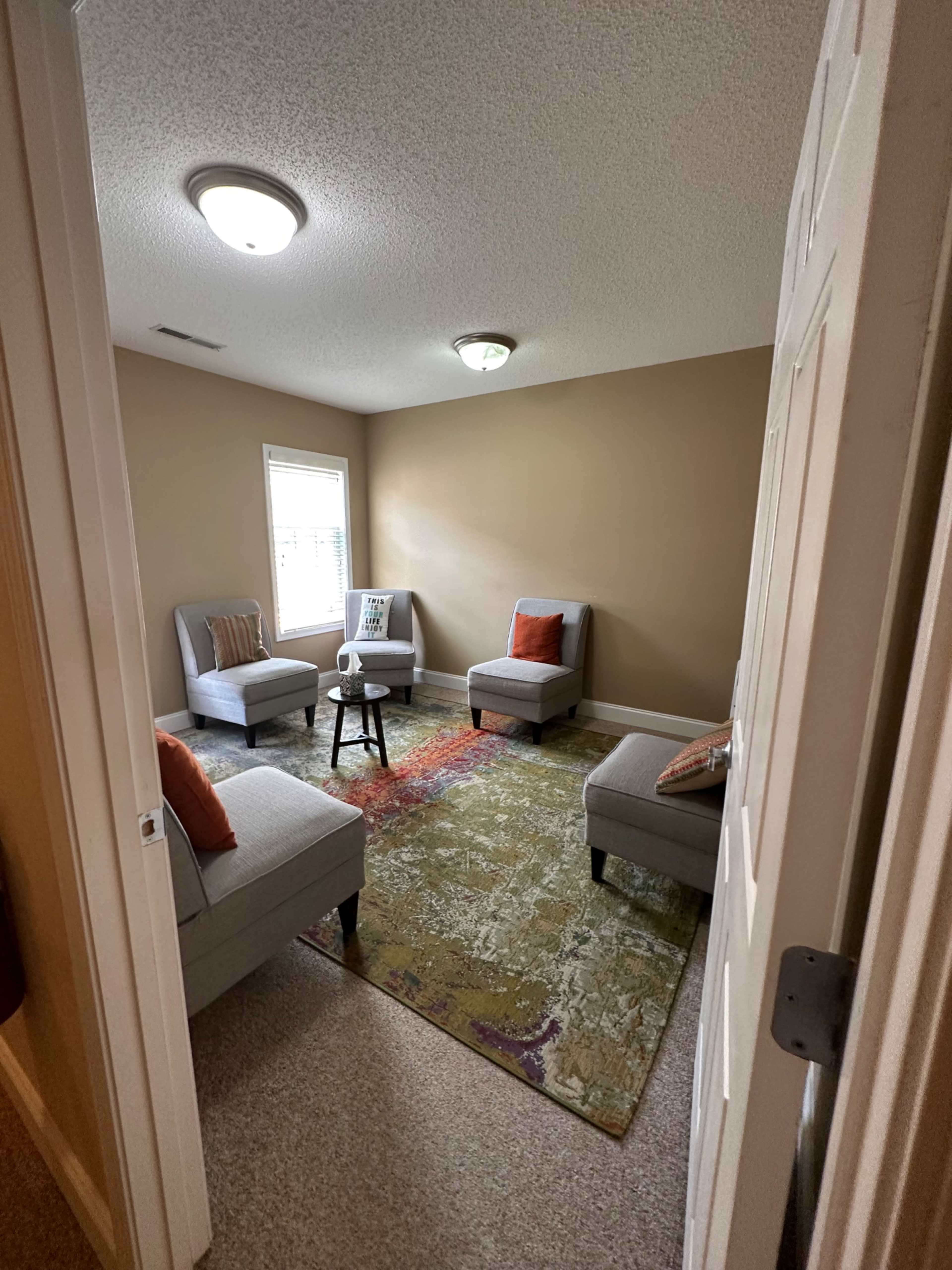 The image shows a small room with four gray chairs arranged around a small black table, with a colorful area rug on the floor and a window allowing natural light.