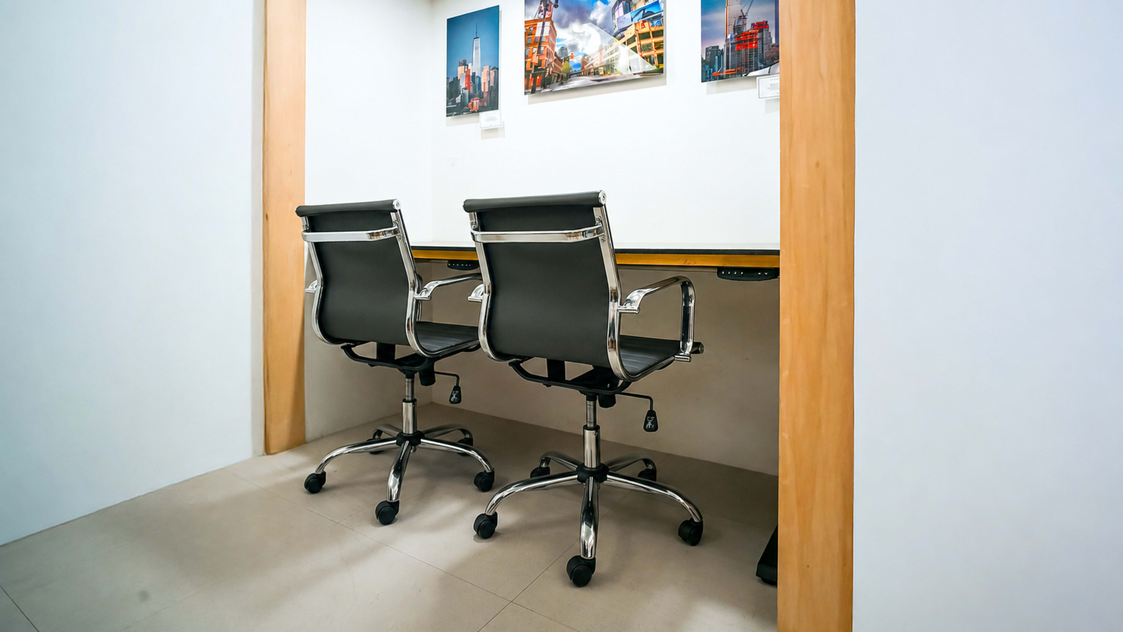 The image shows a minimalist office space with two black ergonomic chairs positioned at a wooden desk against a white wall, featuring framed pictures above.